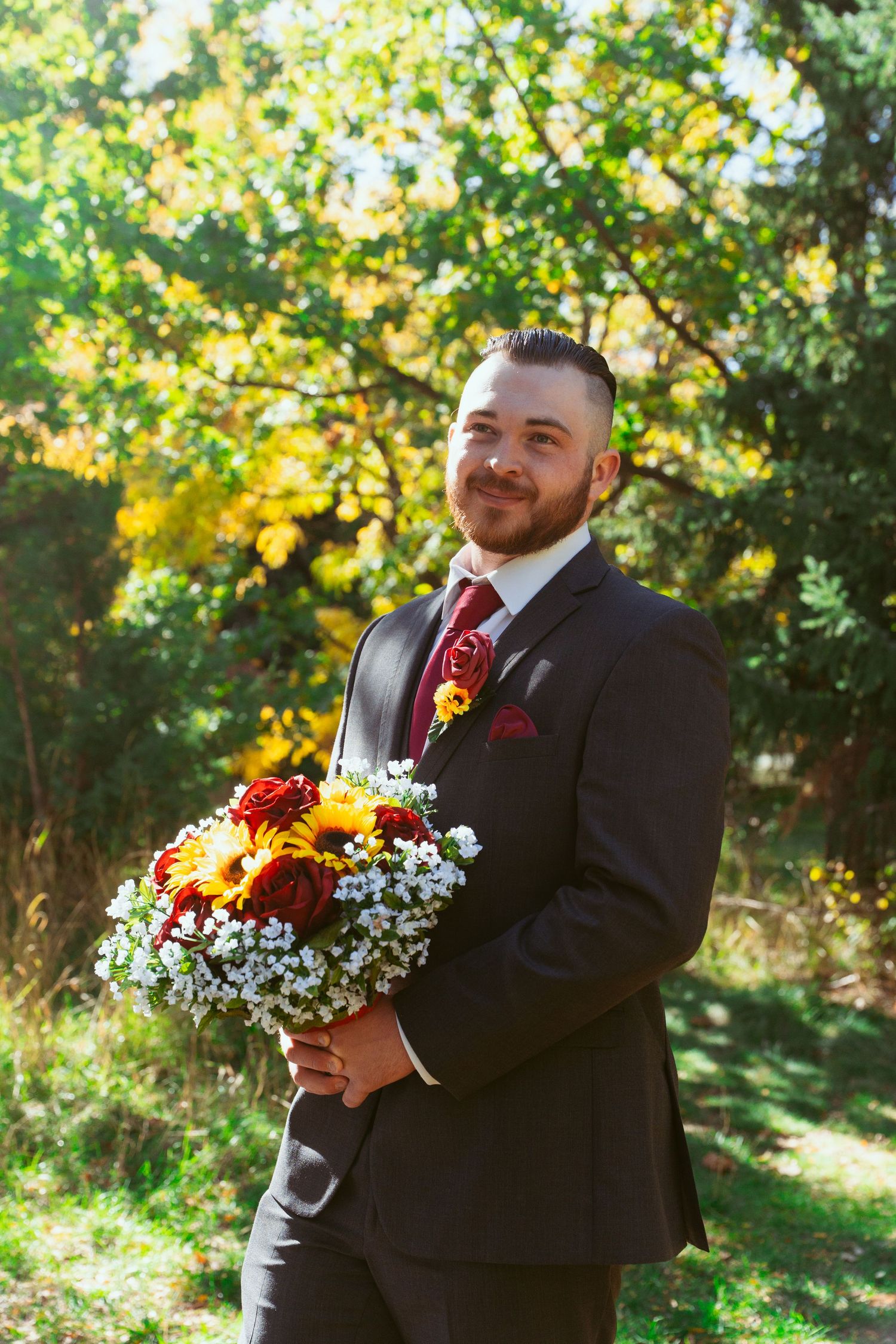 Wedding party members in black suits holding vibrant autumn-colored bouquets in a sunny garden setting.