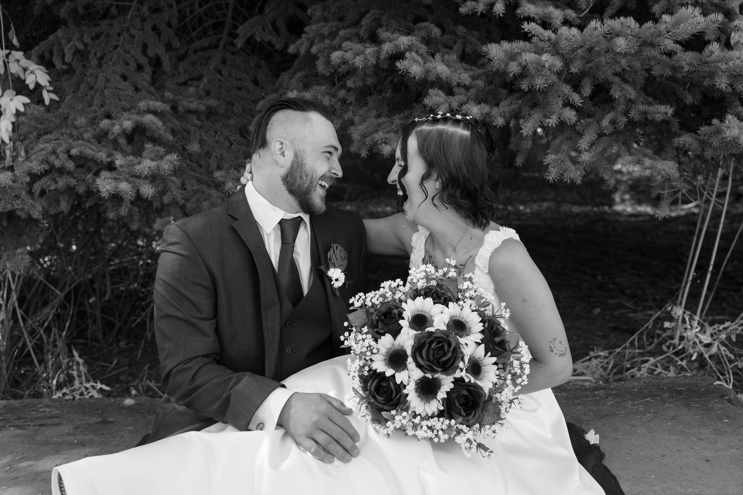 Black and white wedding photo of couple sitting on stone bench in garden setting.