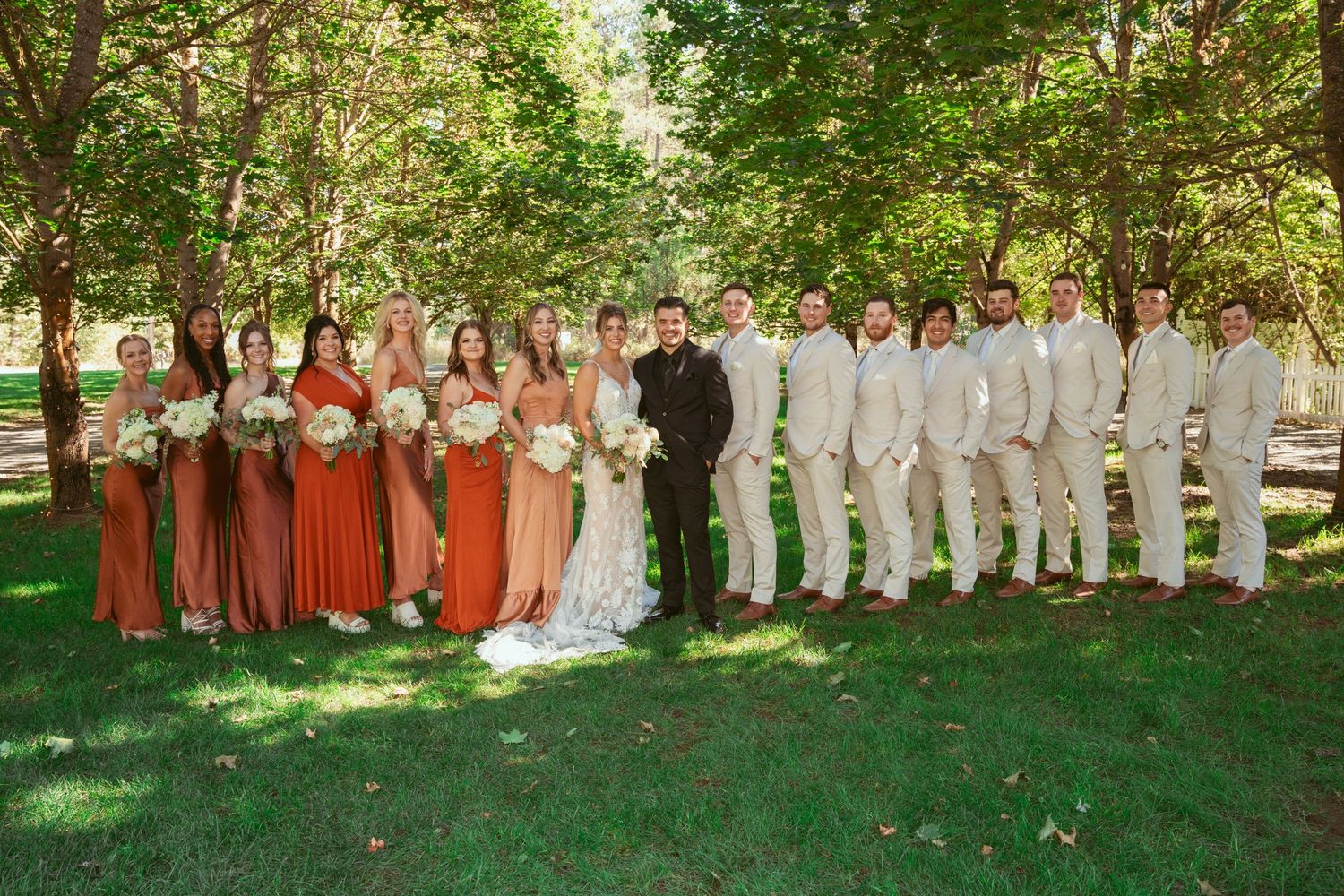 Full wedding party in coordinated formal attire gathers for group photo in outdoor garden setting.