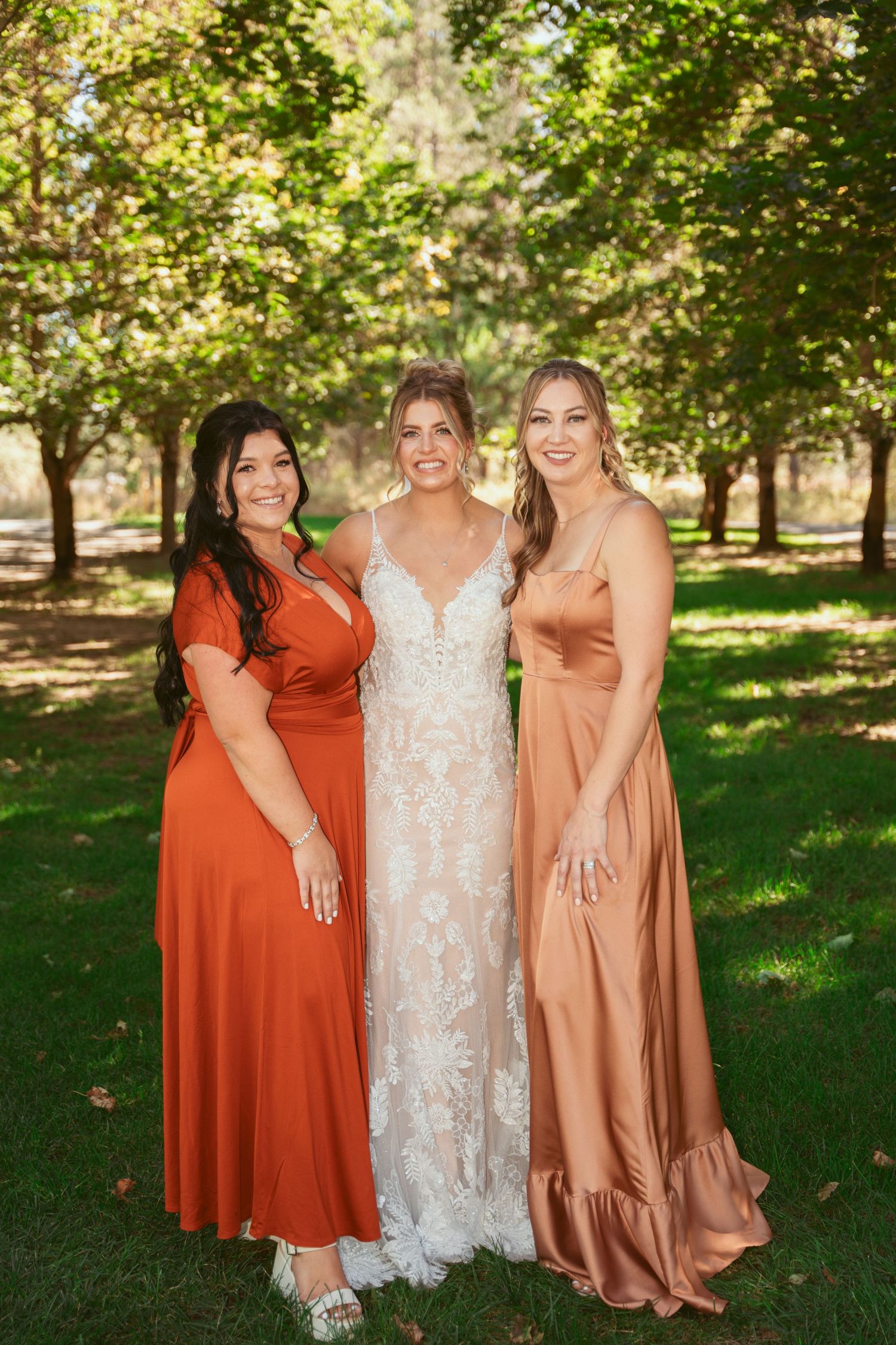 Wedding party poses outdoors in coordinated rust orange dresses and white lace gown in a sunlit garden setting.