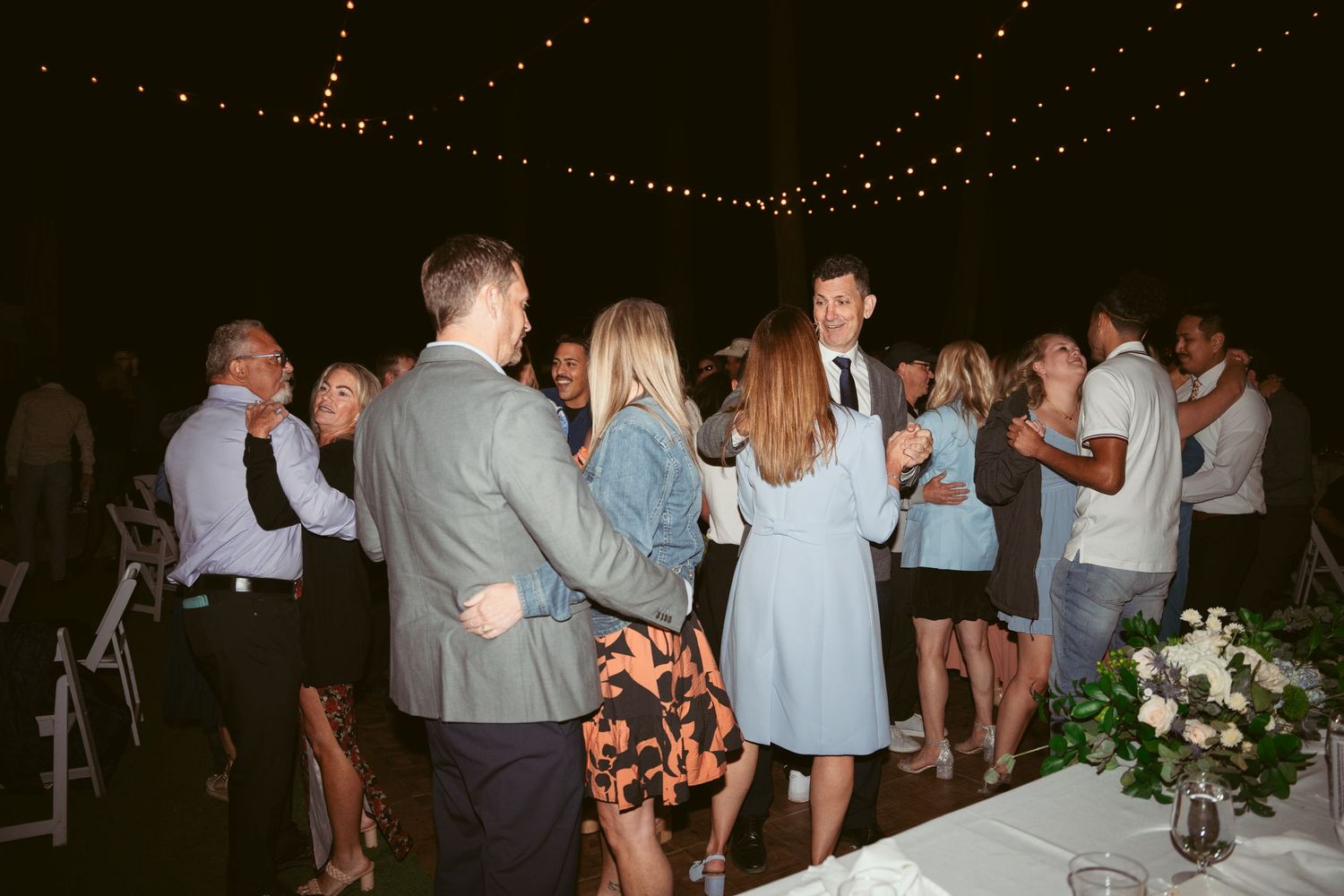 Wedding guests mingling and dancing at an outdoor evening reception under string lights.