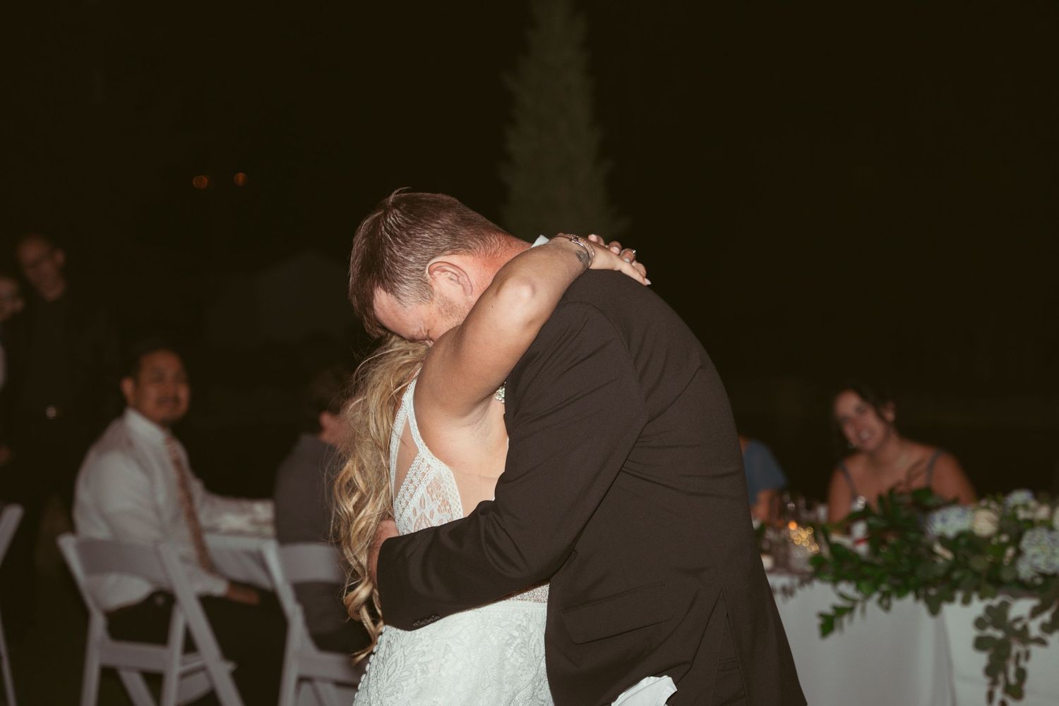 Wedding couple embraces during their reception with decorative lighting in the background.