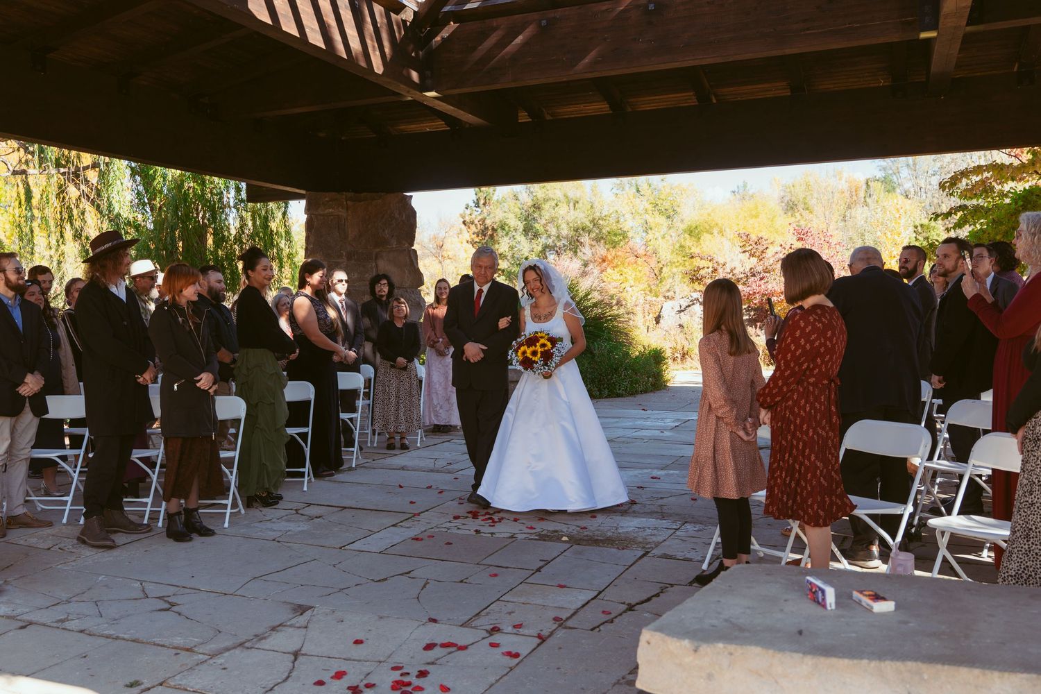 Outdoor wedding ceremony with guests seated on white chairs under covered stone patio.