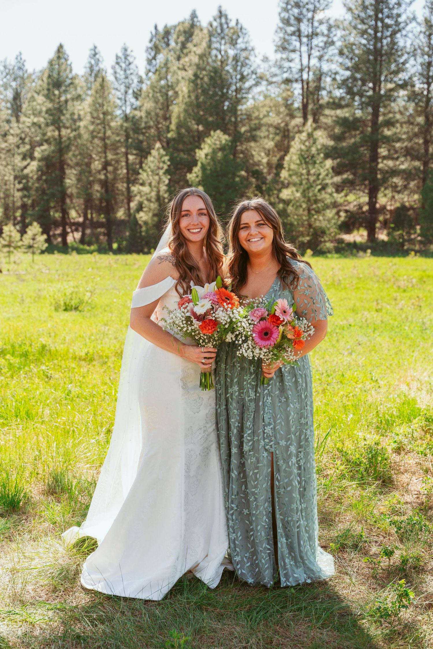 Wedding party members hold bouquets while posing together between tall pine trees.