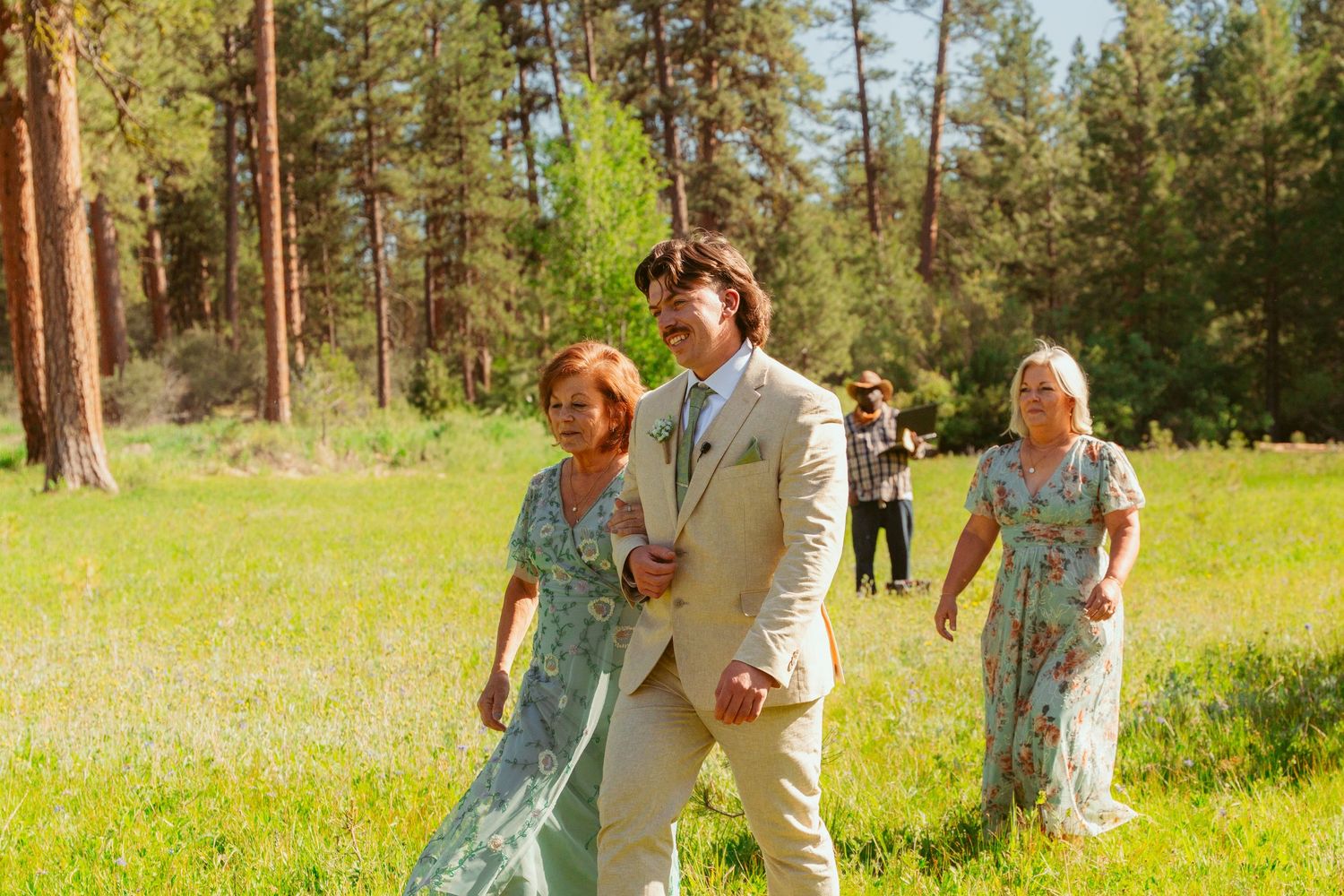 Wedding party and guests celebrate on a sunny meadow with evergreen forest backdrop.
