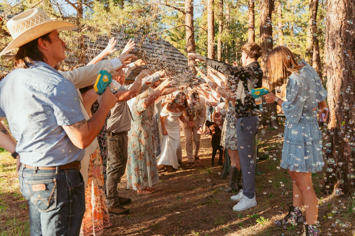Wedding guests spraying celebratory bubbles in sunlit forest setting.