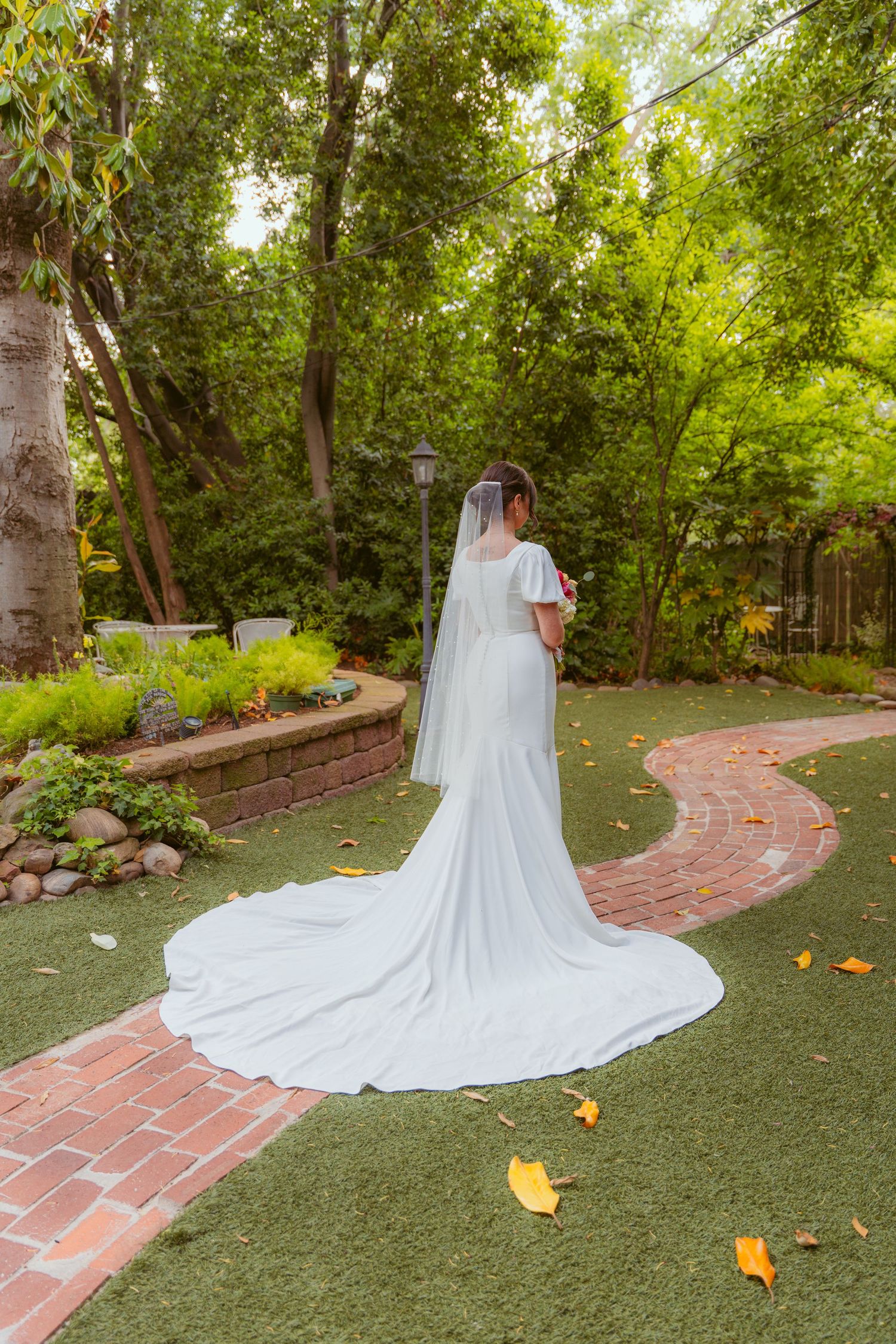 Long white wedding dress train spreads across green lawn with brick pathway and autumn leaves.