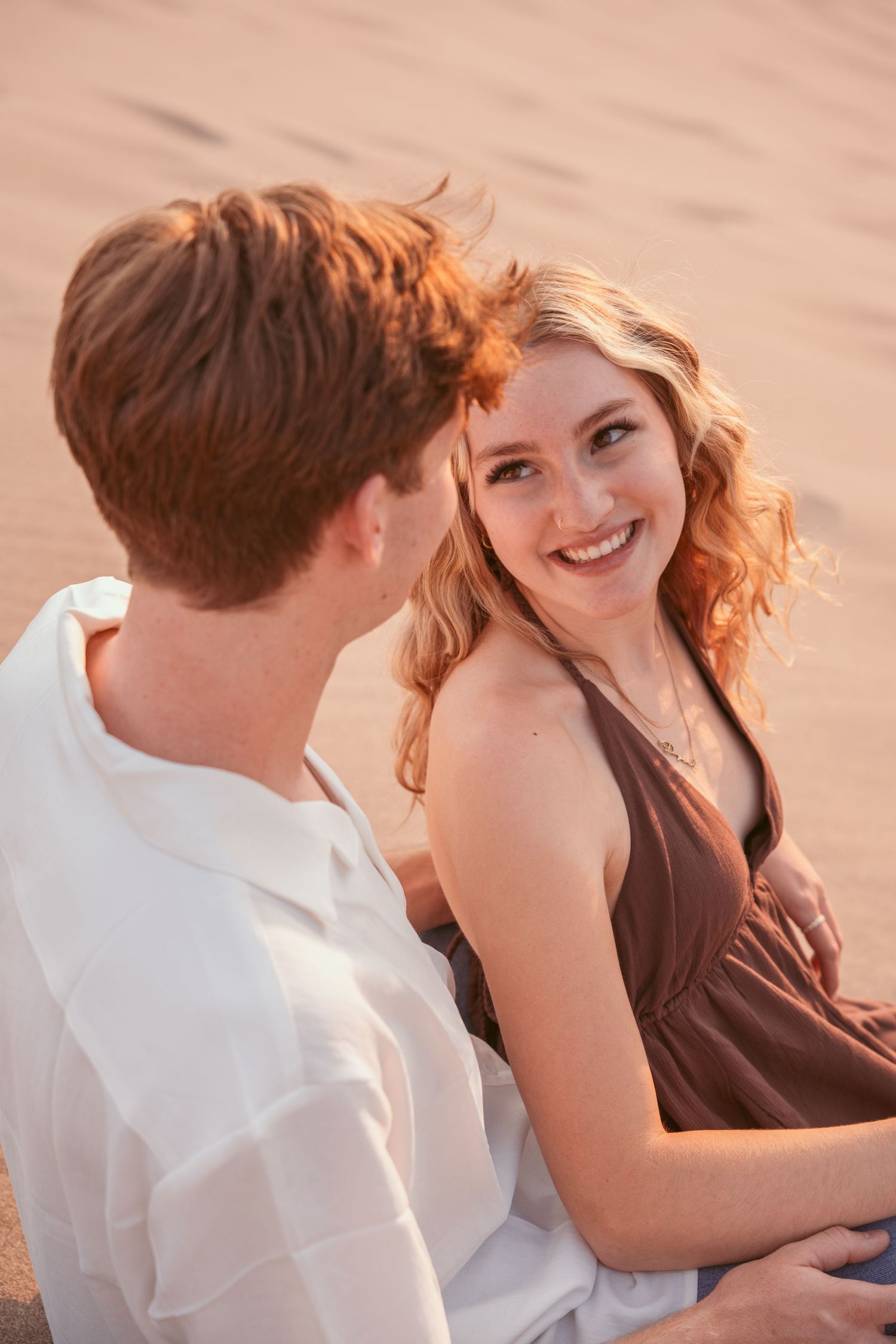 Two people share playful moments together while sitting on sandy dunes at sunset.