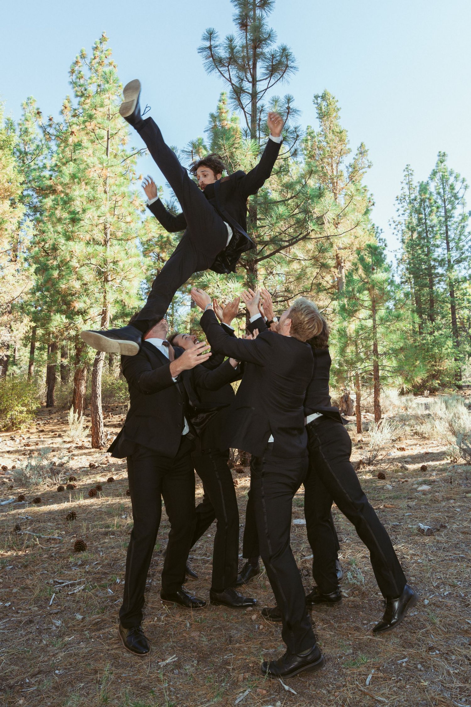 Groomsmen in black suits strike fun poses for group photos in a wooded outdoor setting.