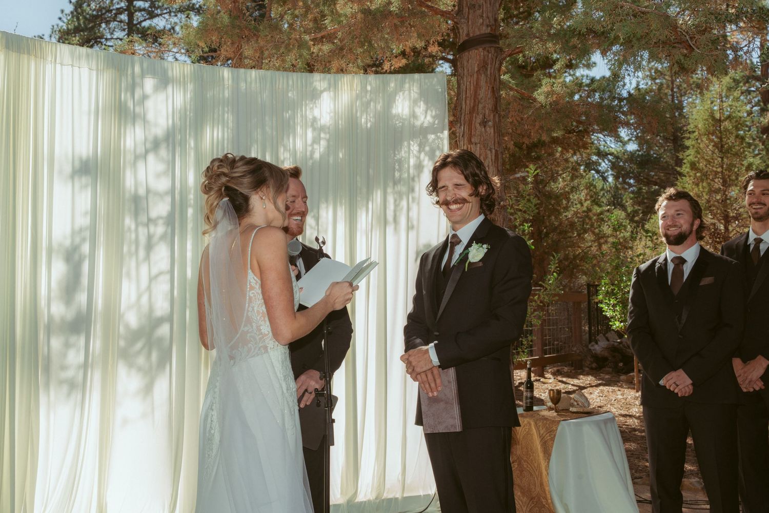 Outdoor wedding ceremony in a forest setting with white fabric backdrop and pine trees.