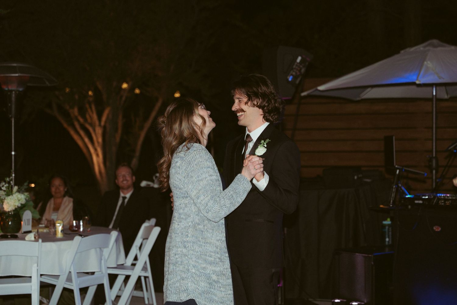 A couple shares their first dance at an outdoor evening wedding reception with white chairs and blue lighting.