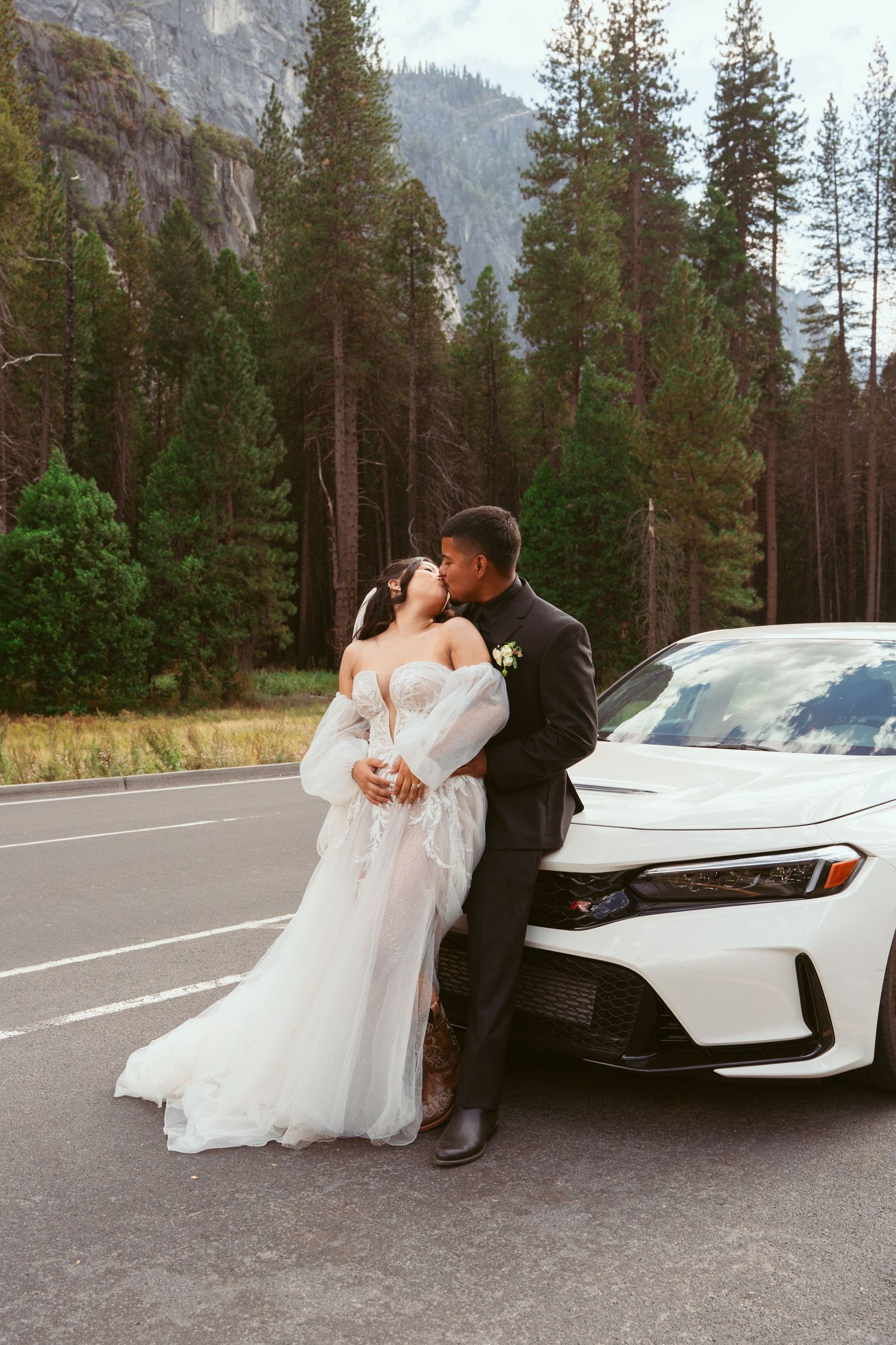 Wedding couple posing romantically next to white car with mountain landscape behind.
