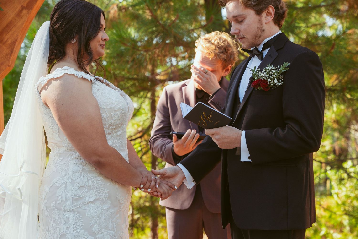 Wedding ceremony moment captured in sunlit forest setting with wooden arch.