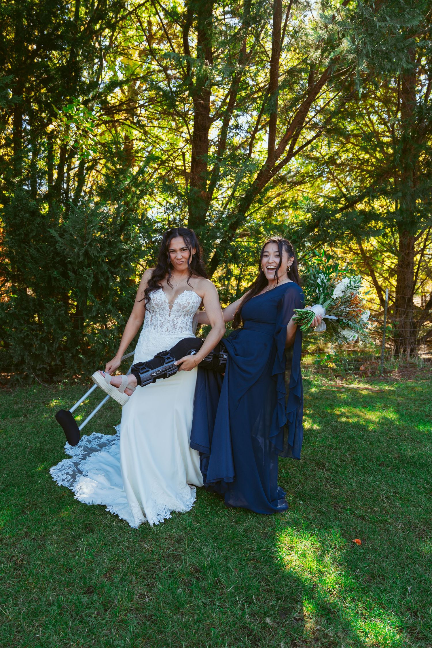 Wedding party members in navy and white dresses sit together on grass under sunlit trees.
