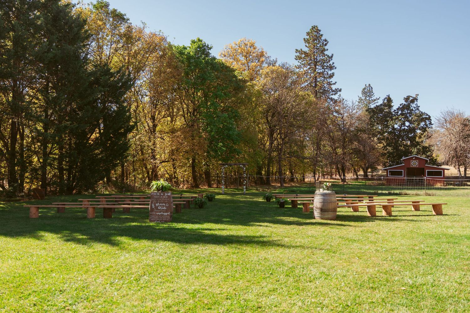 Outdoor ceremony space with wooden benches arranged on sunny lawn surrounded by trees.