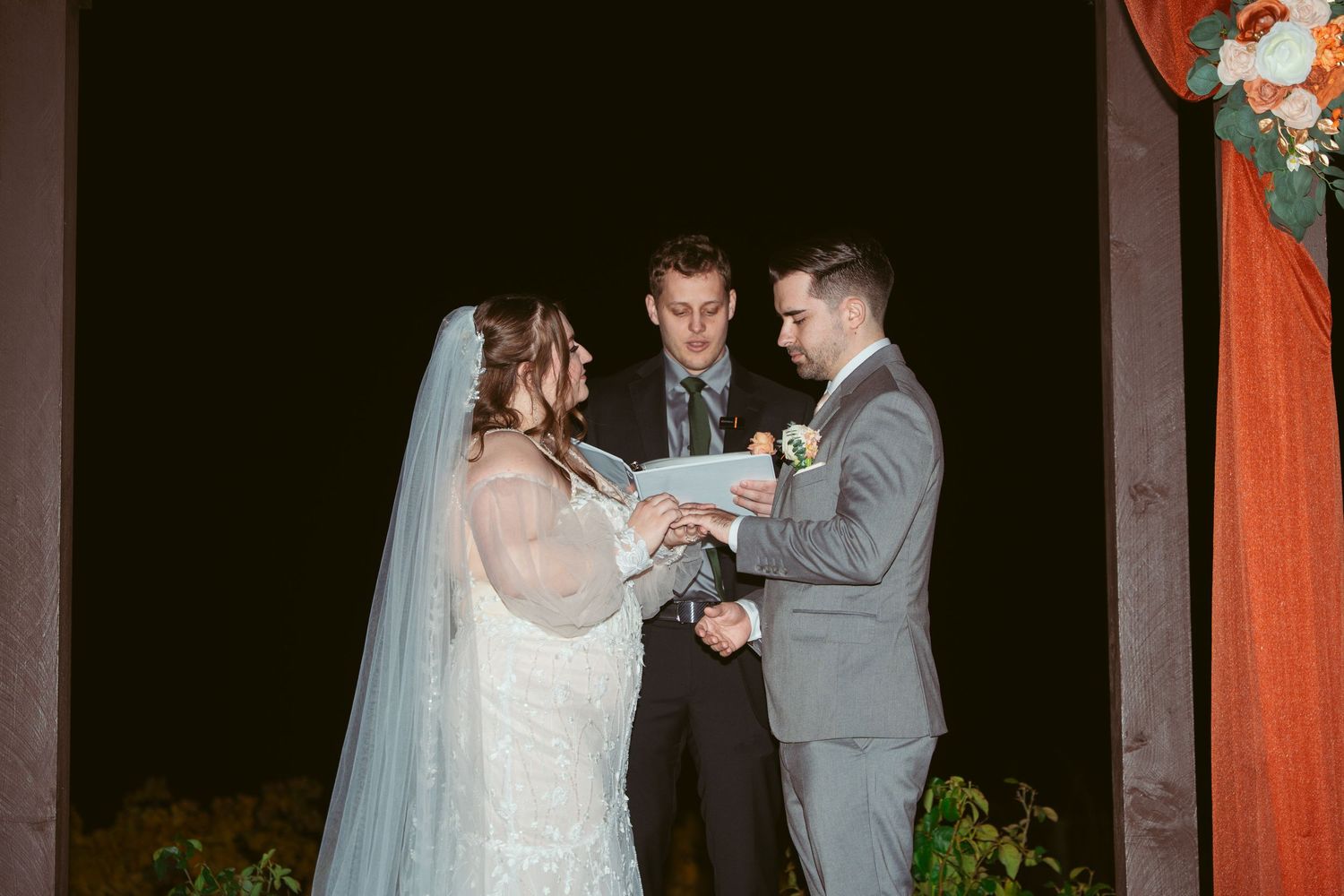 Nighttime outdoor wedding ceremony with couple exchanging vows under illuminated gazebo with orange draping.