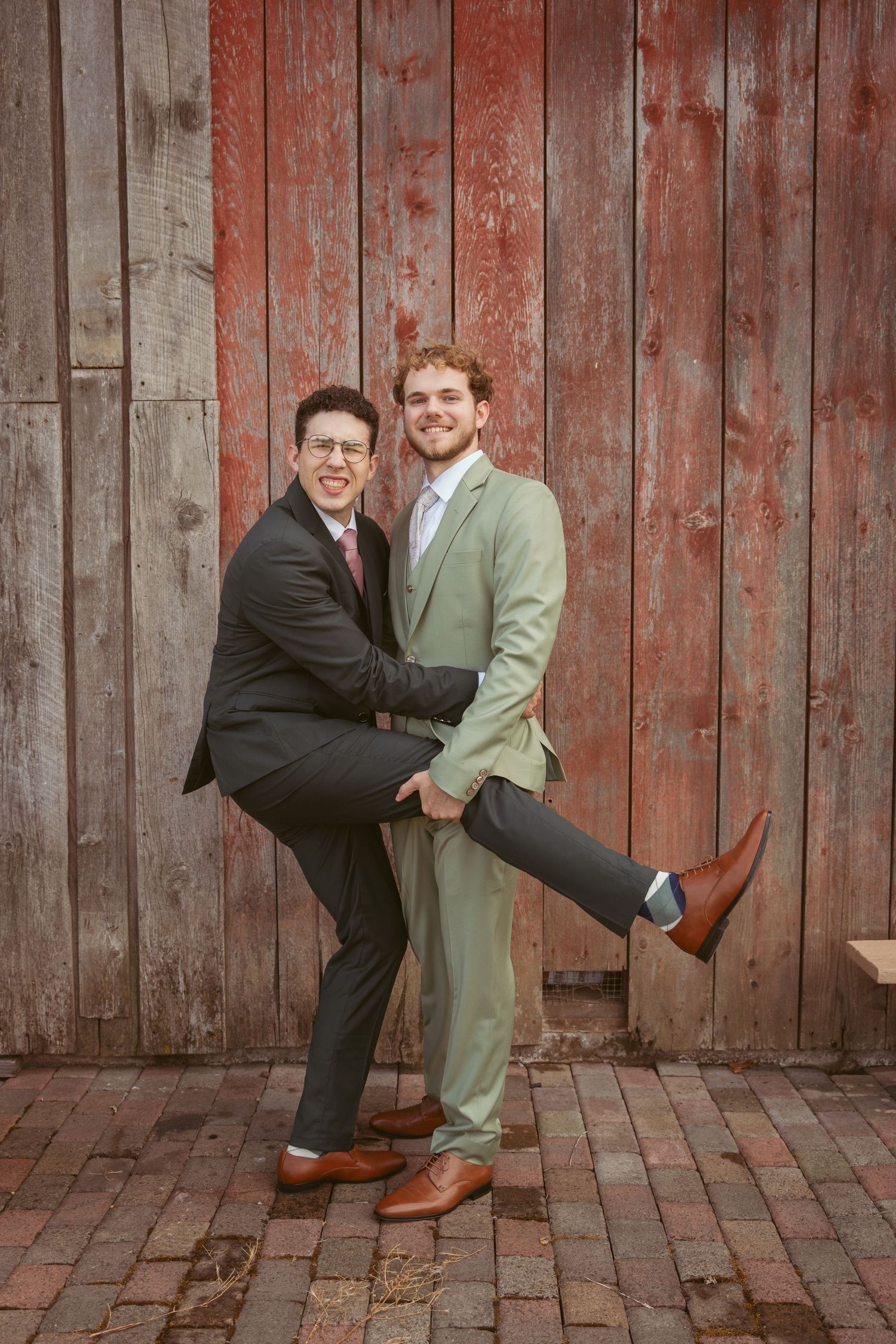 Fun wedding portrait with couple striking playful pose against red barn backdrop.