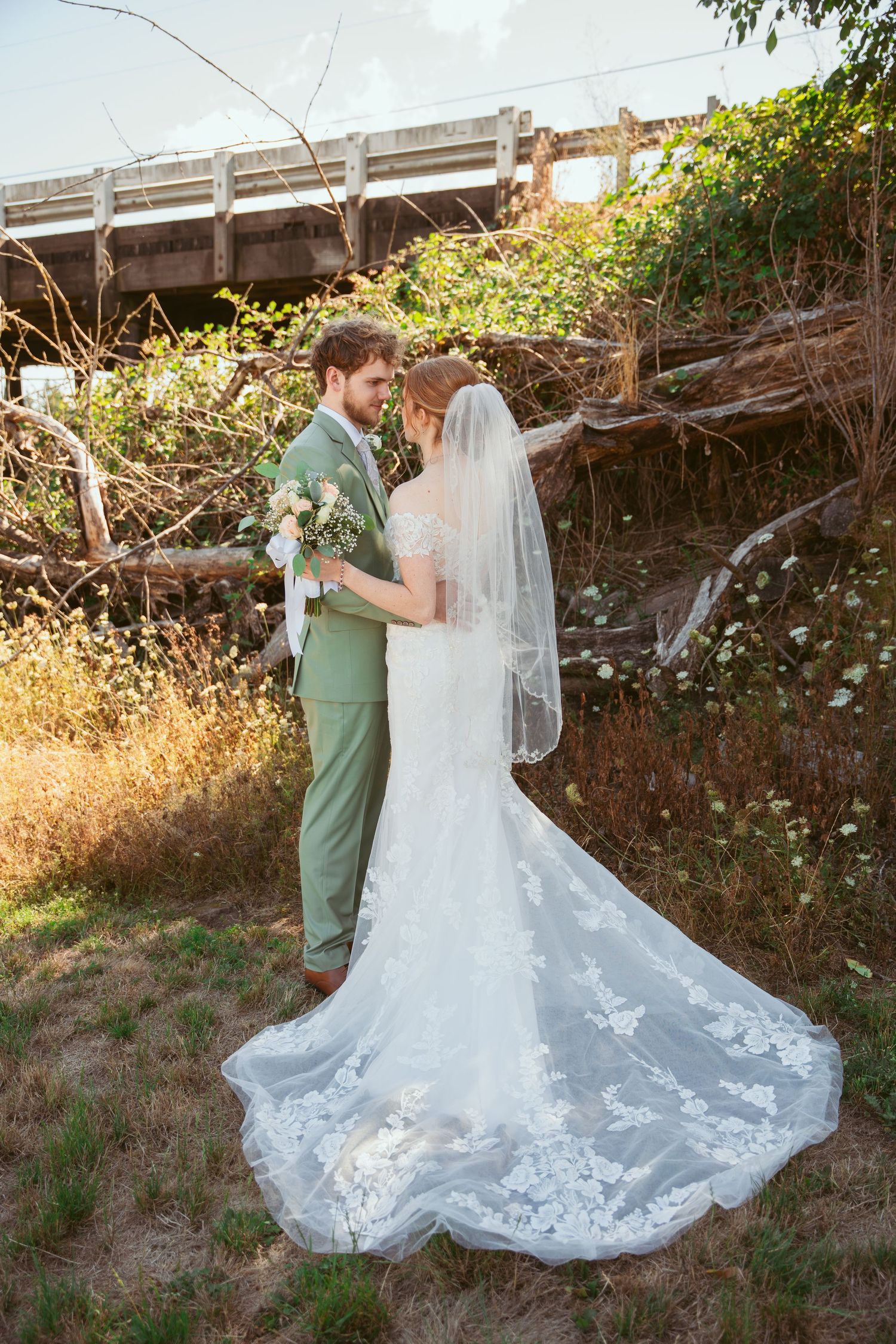 A couple shares an intimate moment outdoors, with the bride wearing a flowing lace wedding dress with a long train.
