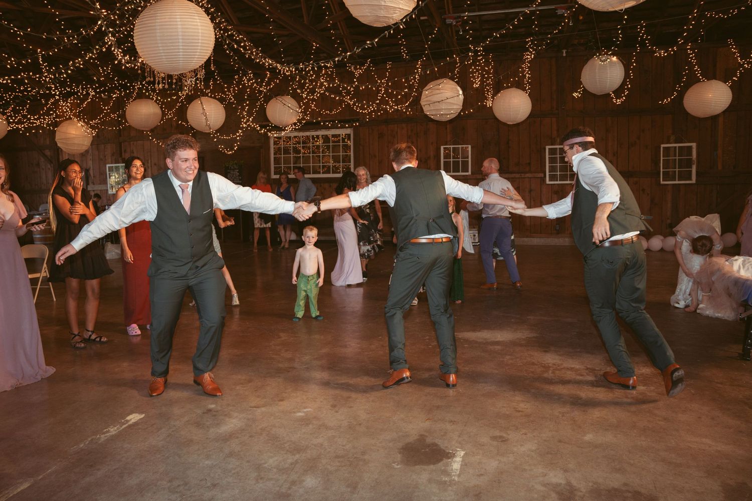 Wedding guests line dancing in a barn decorated with string lights and paper lanterns.