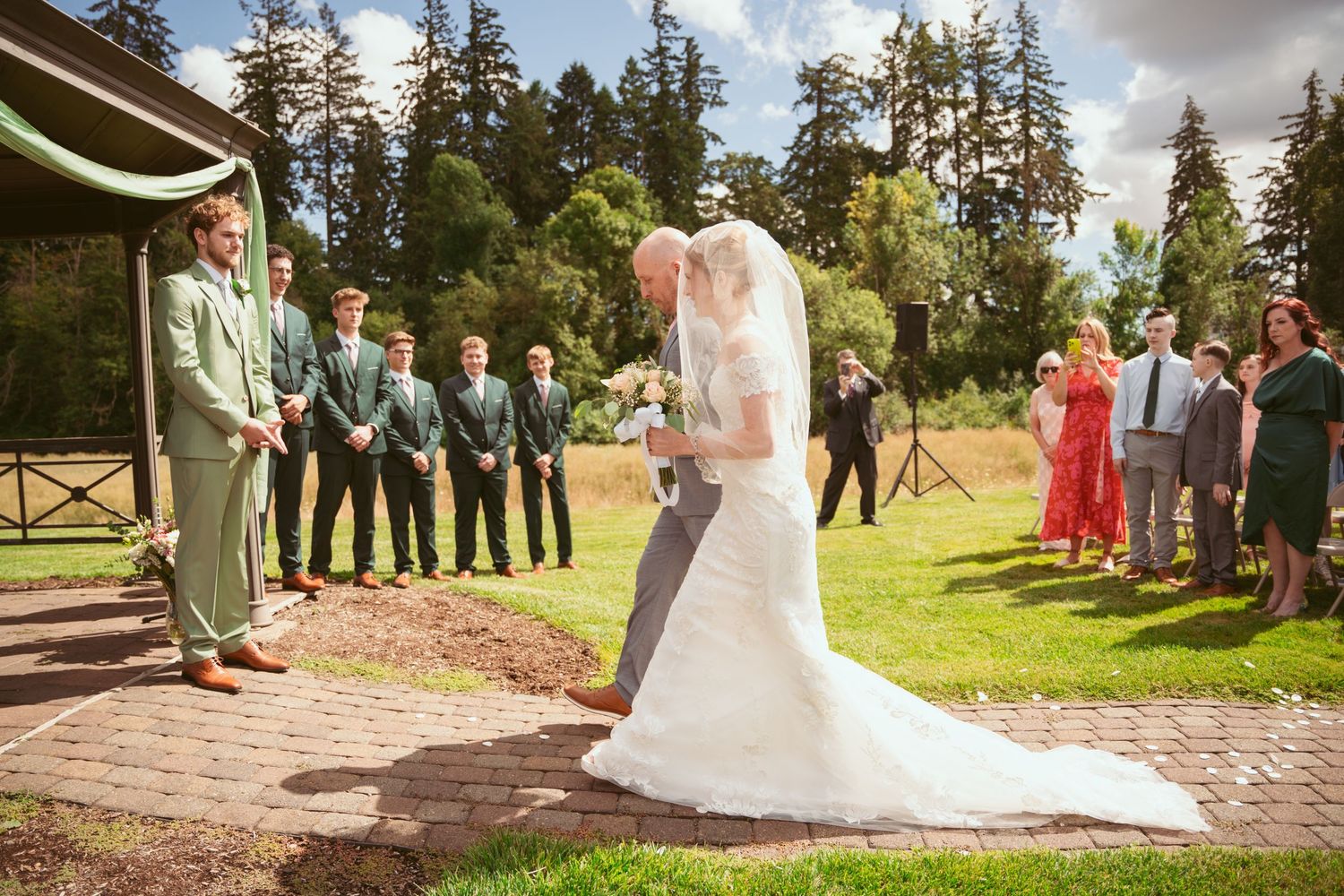Wedding ceremony with wedding party gathered under wooden gazebo on brick pathway.