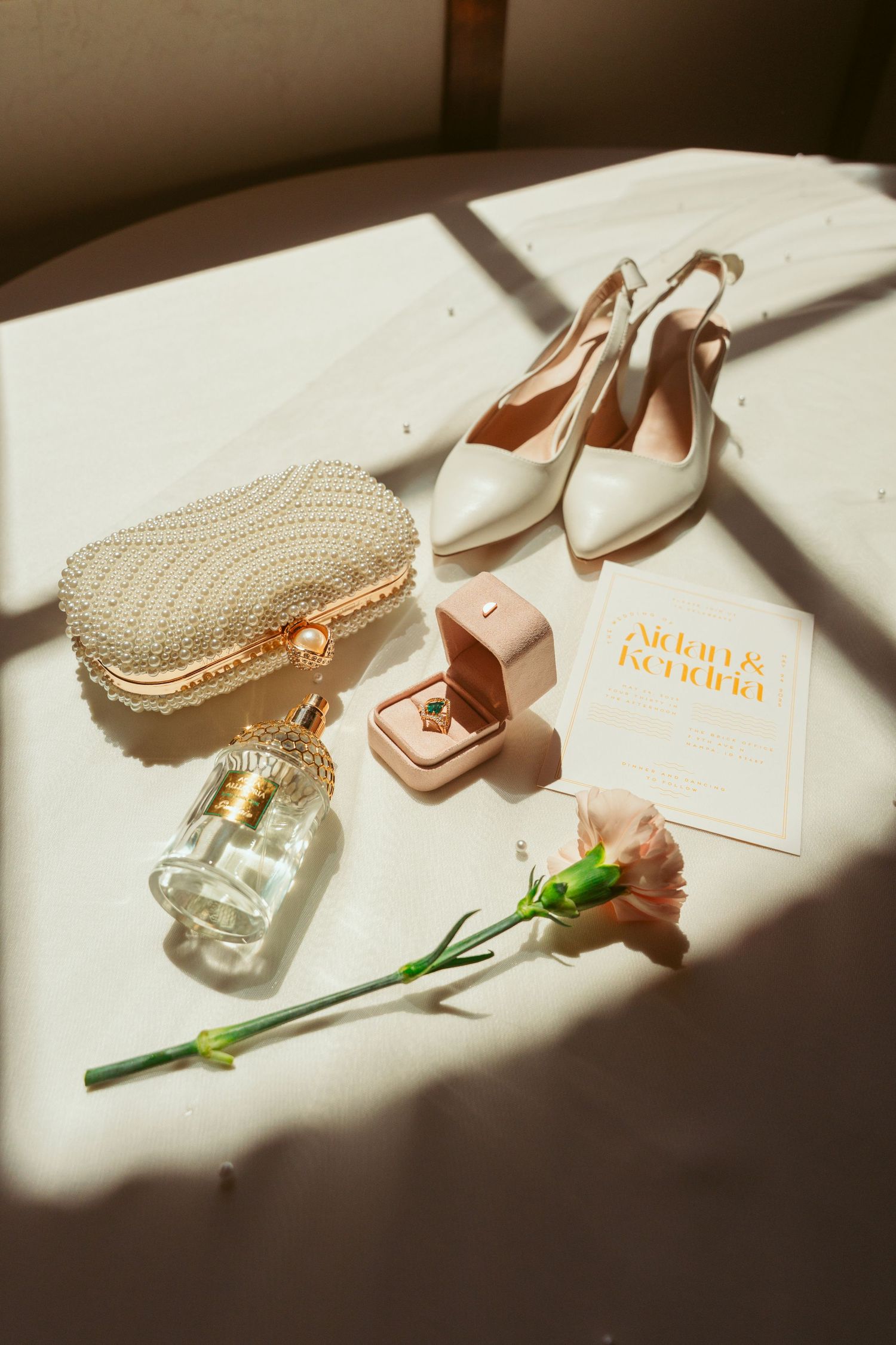Wedding details with white heels, perfume bottle, ring box and invitation in sunlight.