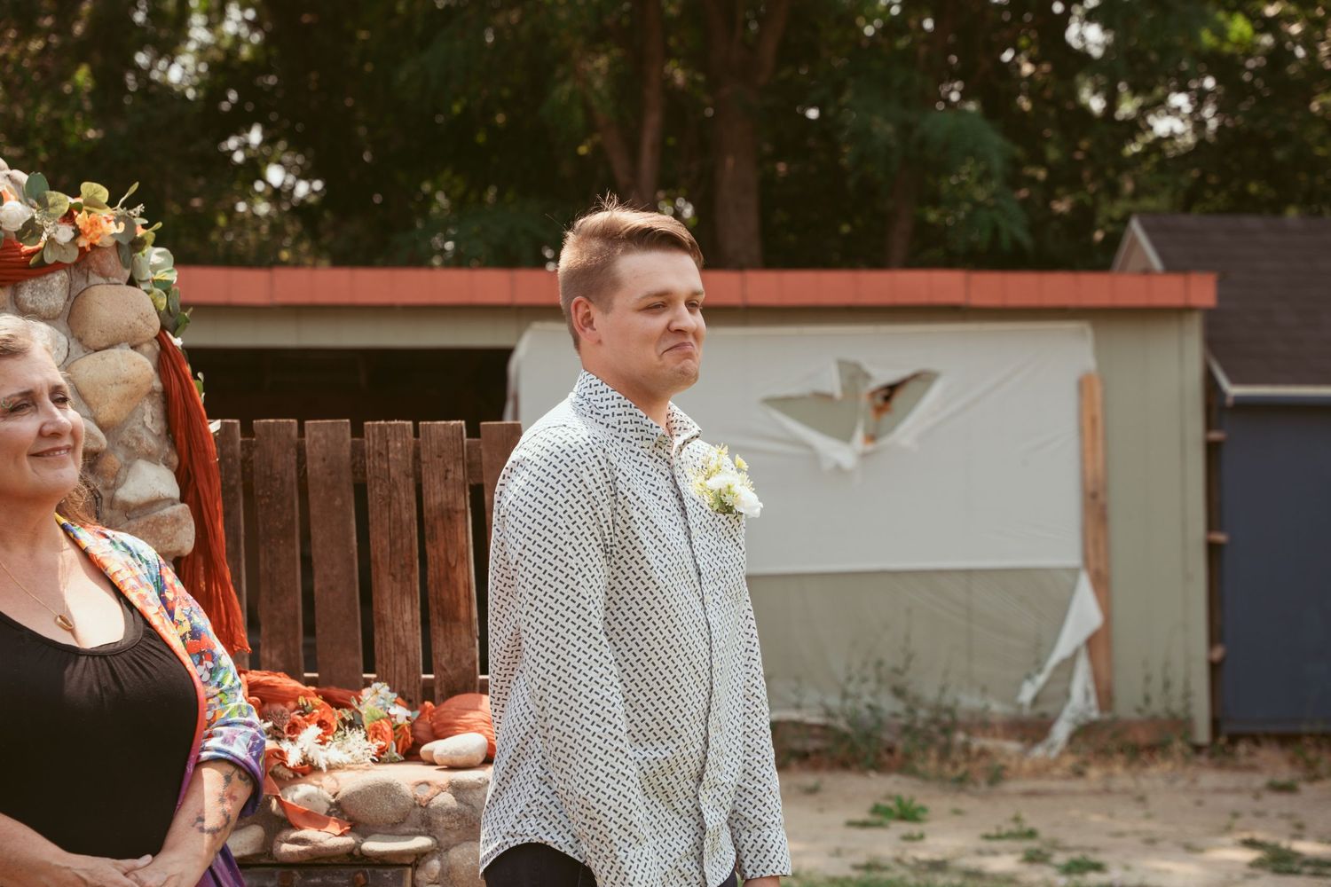 A groom in a patterned shirt stands on a wooden deck during an outdoor ceremony.