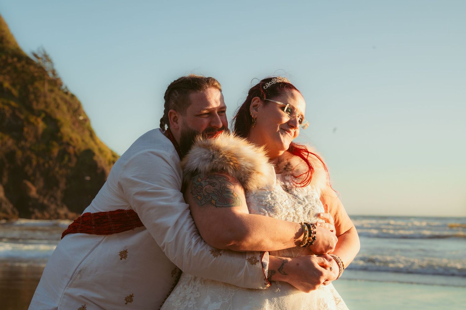 A couple in white wedding attire embraces on a beach with ocean waves and cliffs in the background.
