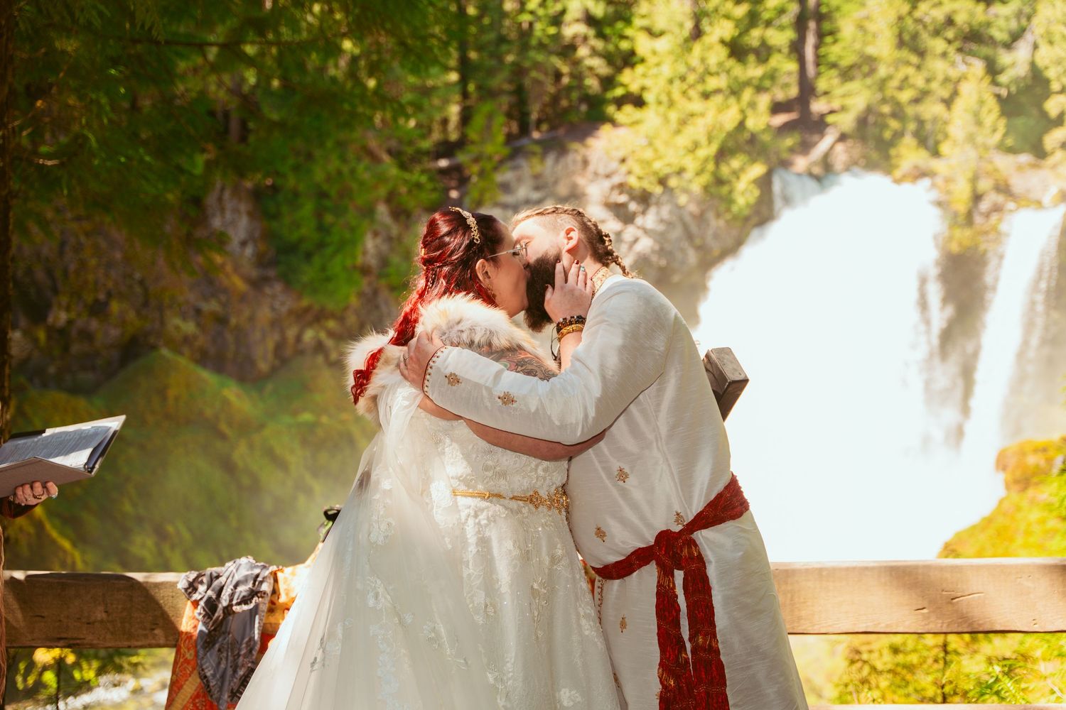 A romantic wedding ceremony takes place in front of a cascading waterfall in a forest setting.