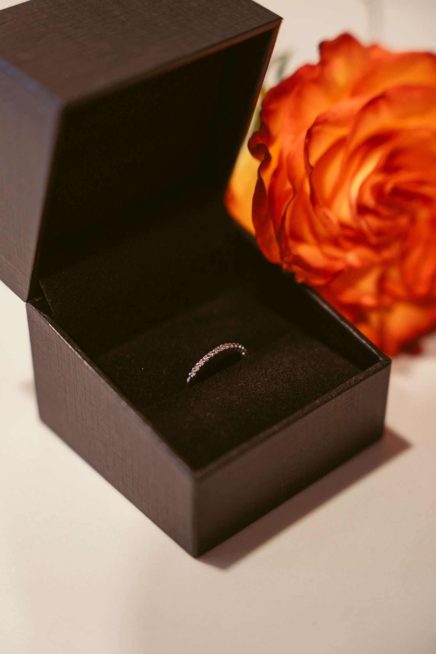 Diamond ring nestled in a black velvet box with an orange rose beside it.
