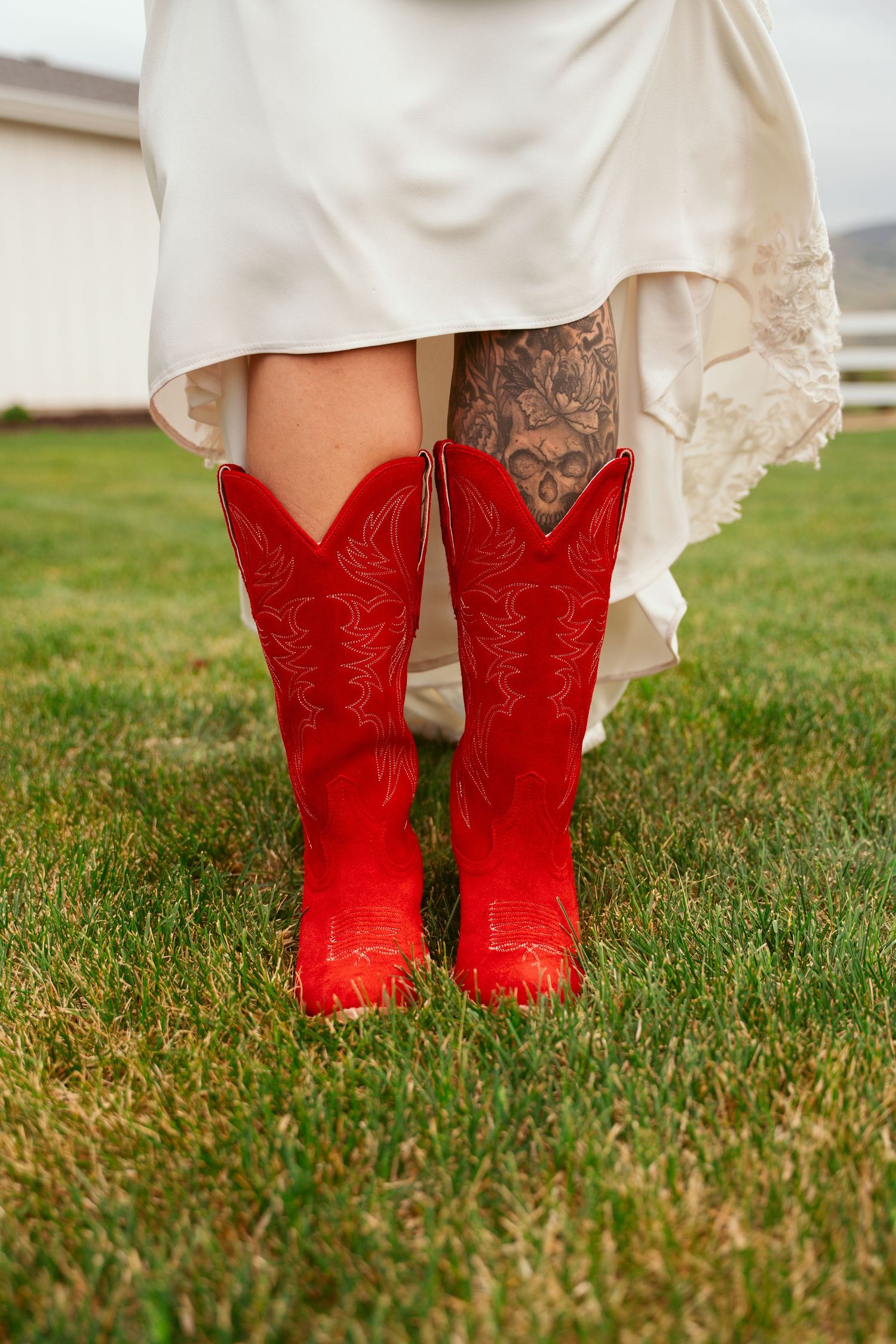Red cowboy boots peeking out under a white wedding dress on grass.