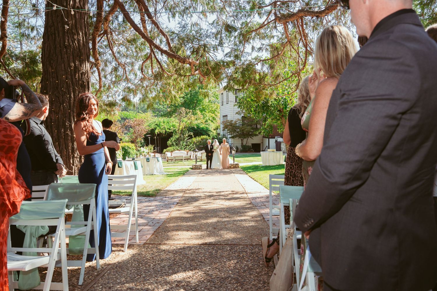 Wedding guests seated in white chairs along a rustic outdoor path surrounded by trees.