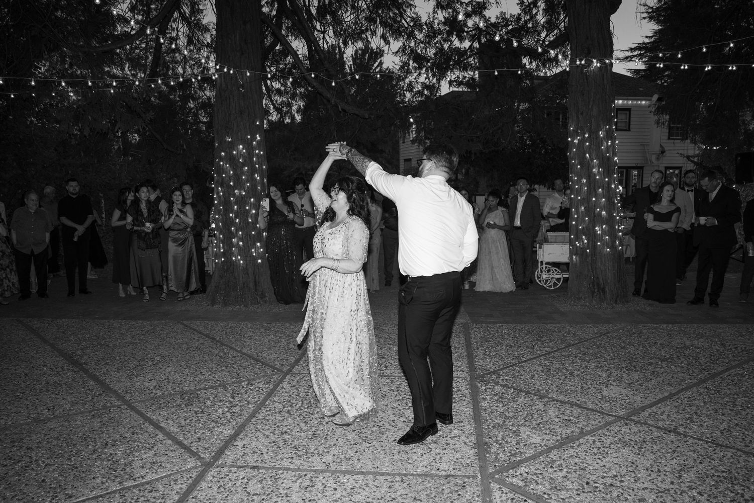 Black and white photo of couples dancing at an outdoor wedding reception under trees.