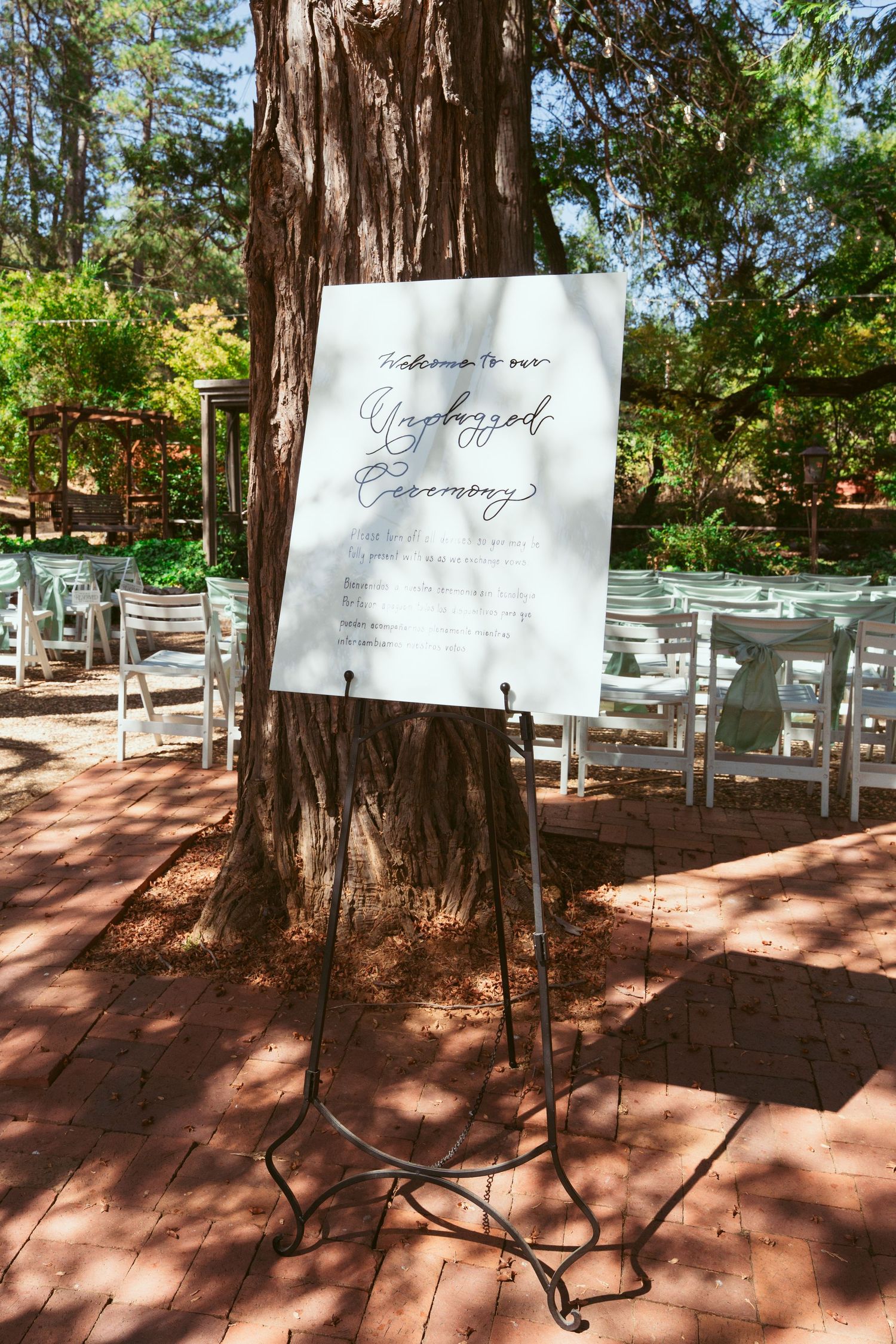 Wedding ceremony welcome sign displayed on an easel stand among outdoor chairs in a wooded setting.