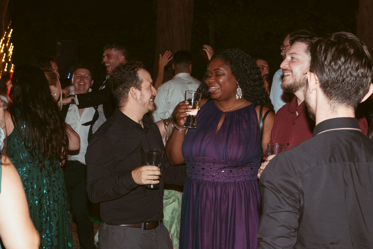 Wedding guests dancing and socializing at a reception with people wearing formal attire and purple dresses.