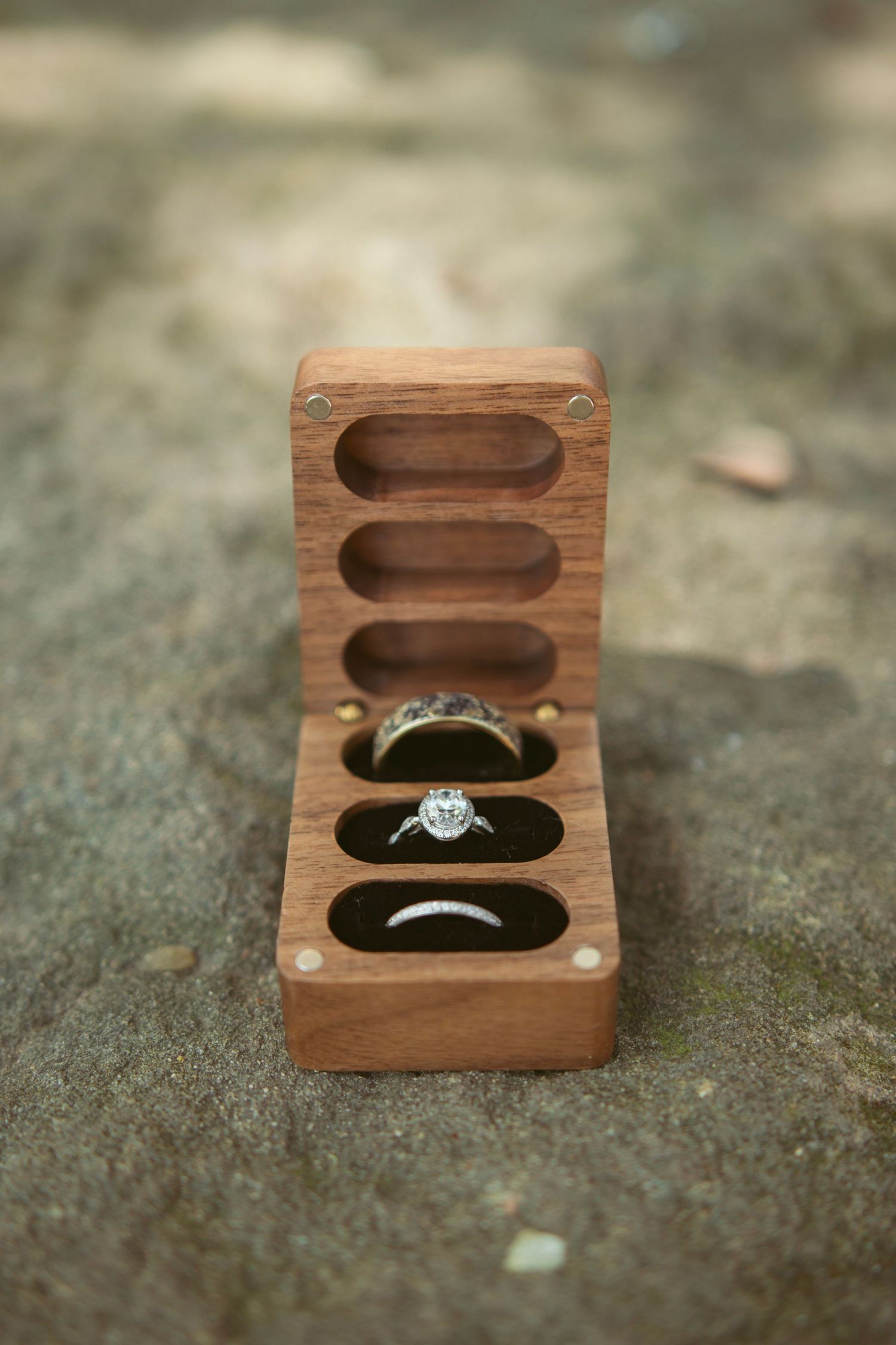 A wooden ring box with a diamond engagement ring shown from multiple angles on a textured stone surface.