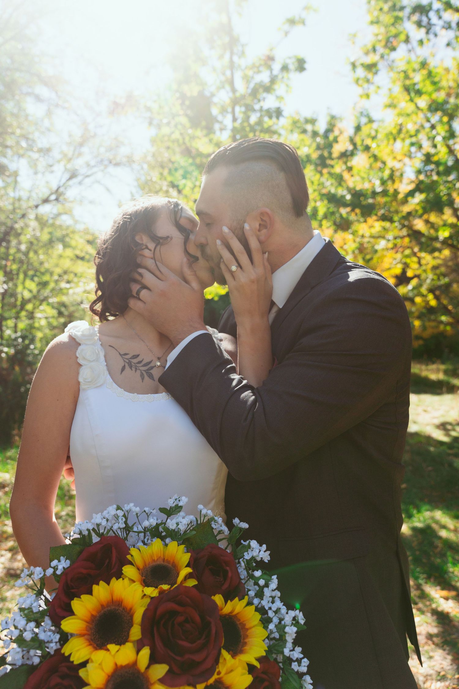 A sun-drenched wedding photo with the couple kissing while holding a vibrant sunflower bouquet.