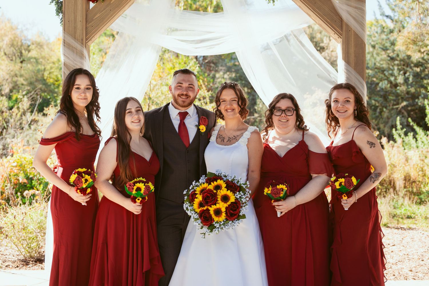 Wedding group photo with bridesmaids in burgundy dresses holding sunflower bouquets.