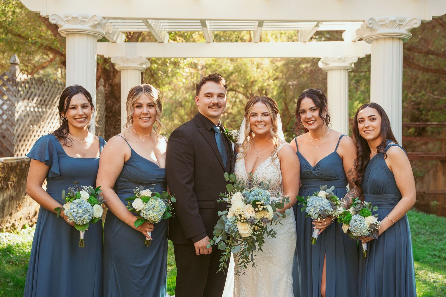 Wedding party in dusty blue dresses poses with couple under white columned pergola with matching bouquets.