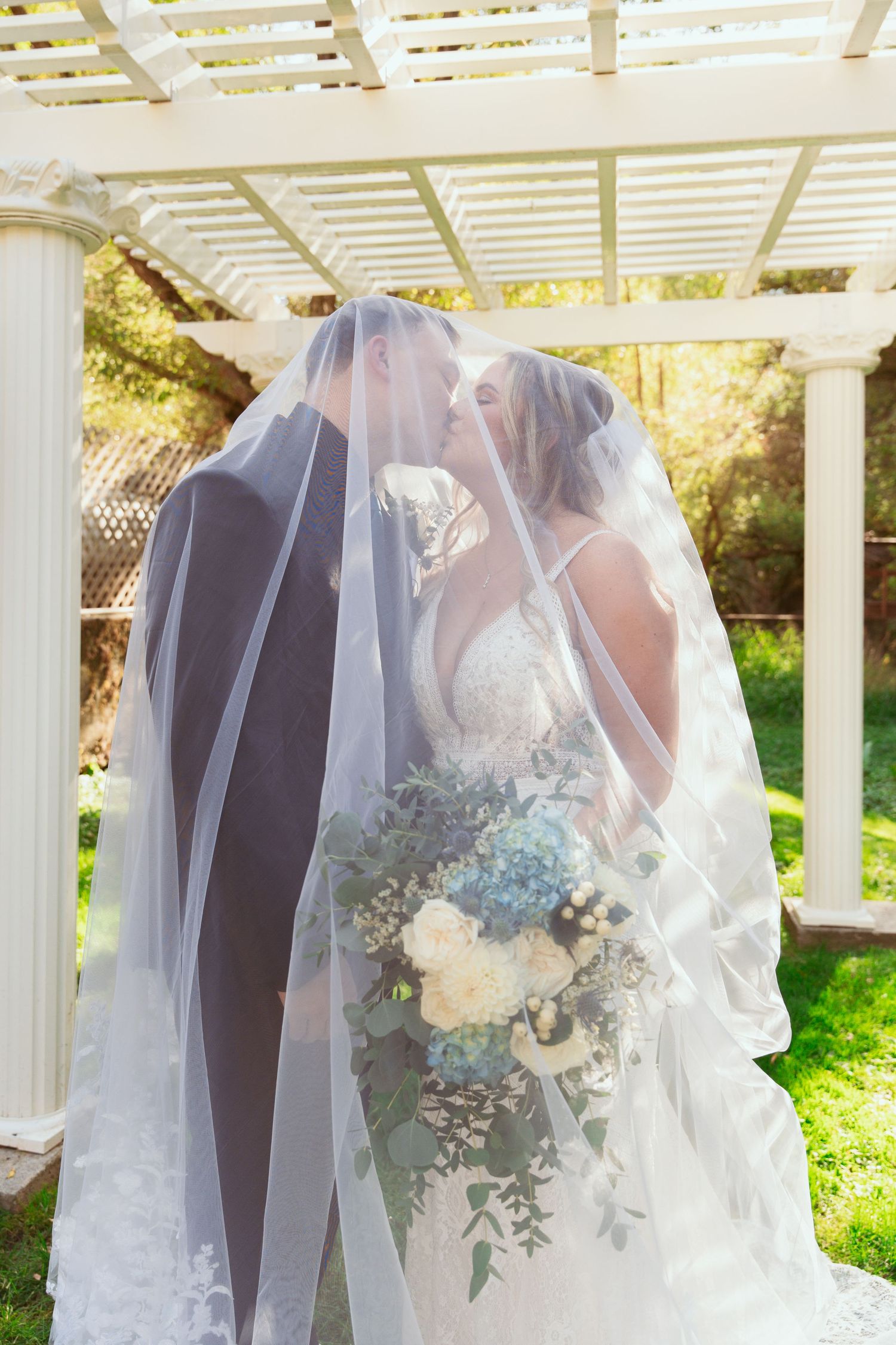 Wedding couple shares intimate moment under white pergola with flowing veil and blue and white floral bouquet.