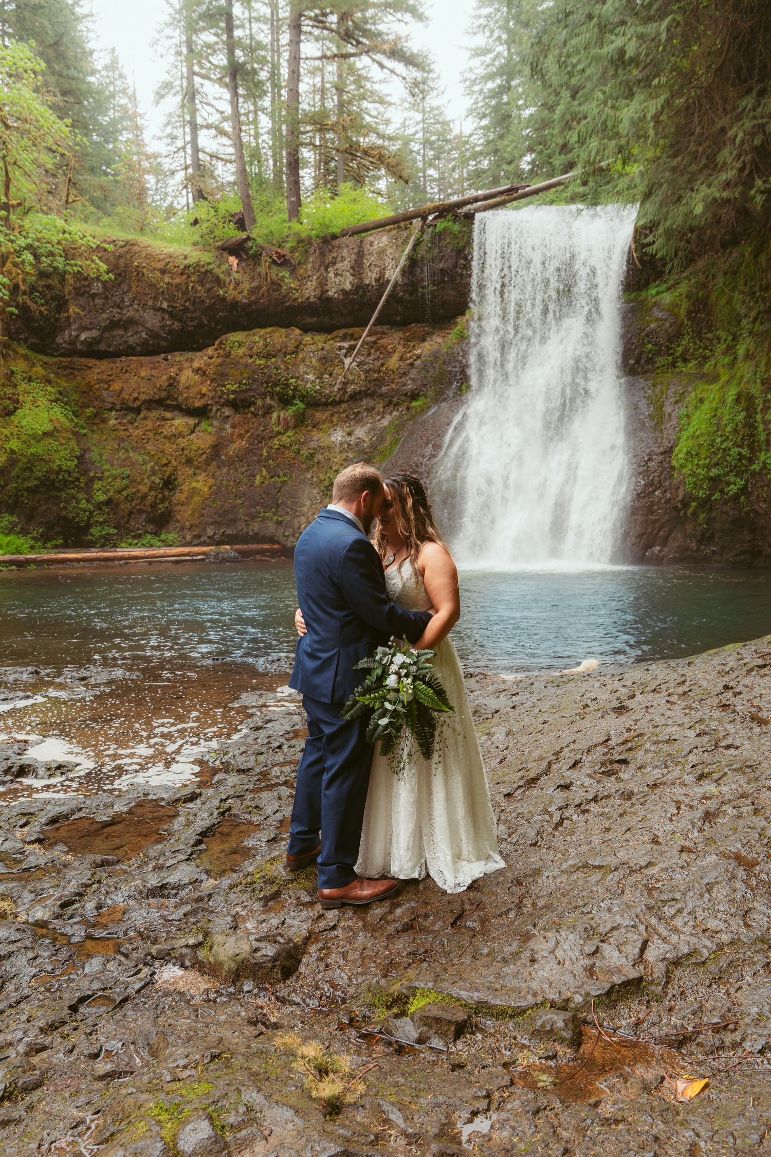 Wedding couple poses on rocky terrain near a flowing waterfall in a peaceful forest setting.