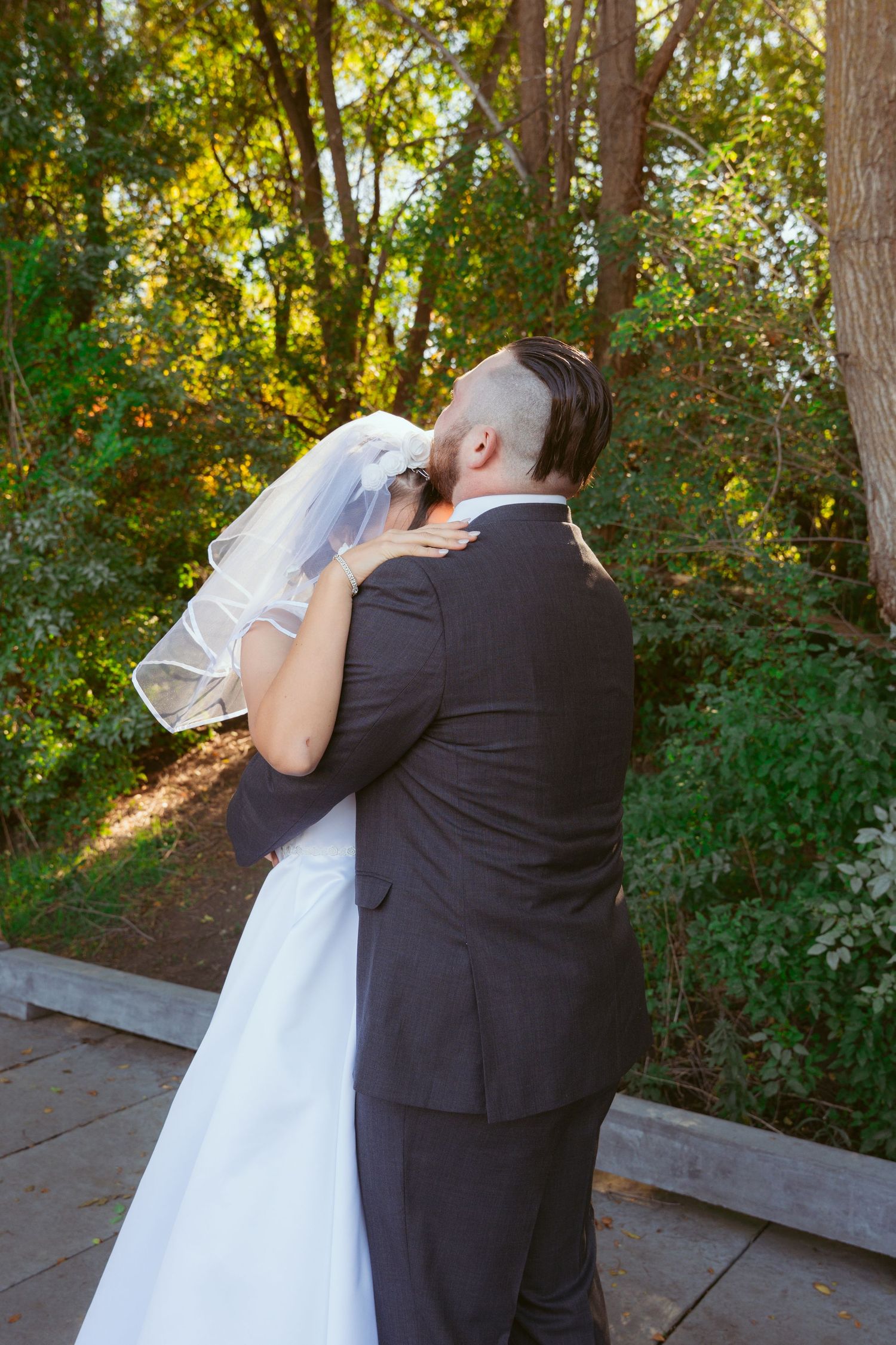 A couple shares a romantic moment on a wooden deck surrounded by lush greenery and sunlit trees.