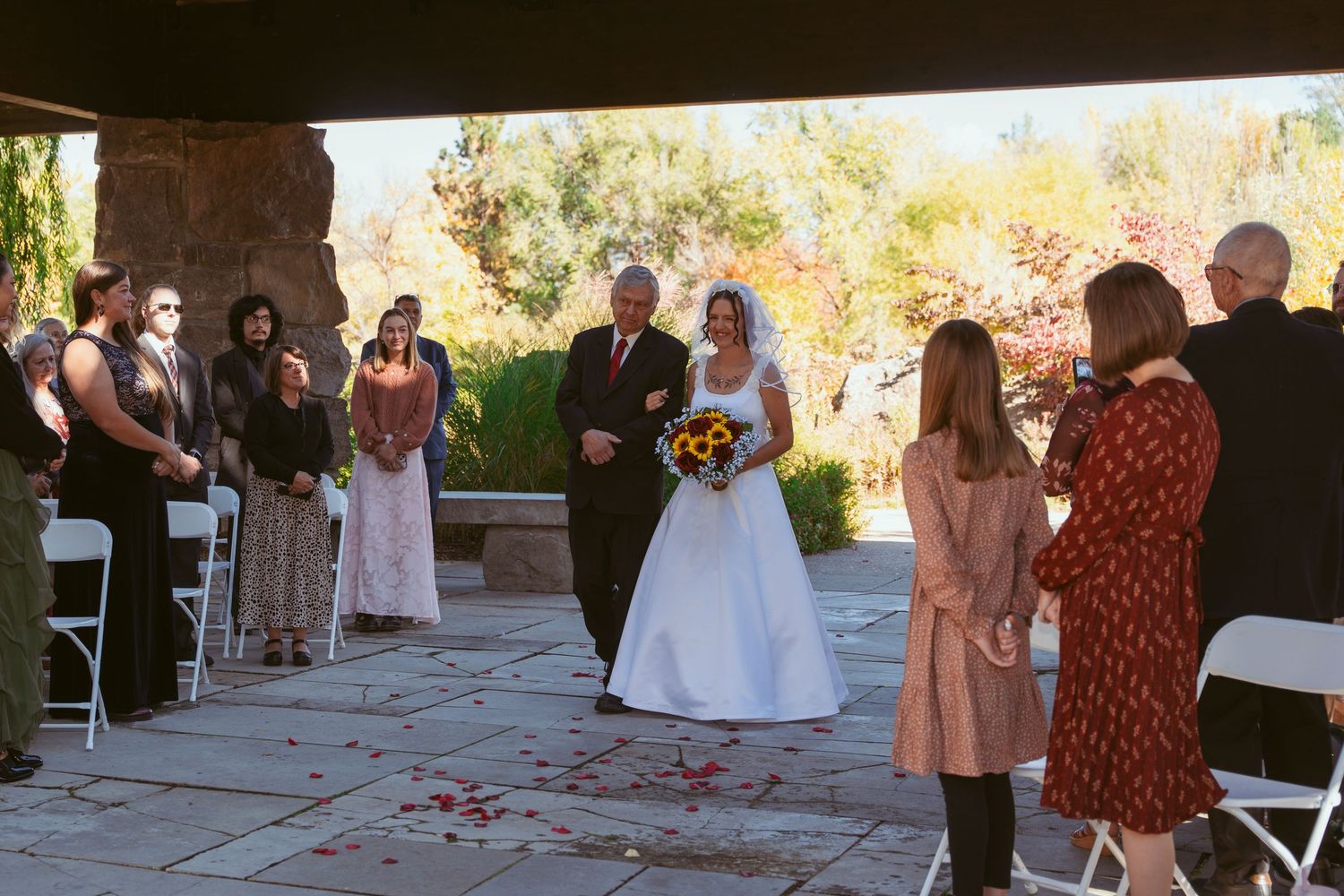 Wedding ceremony processional under covered patio with autumn foliage in background.