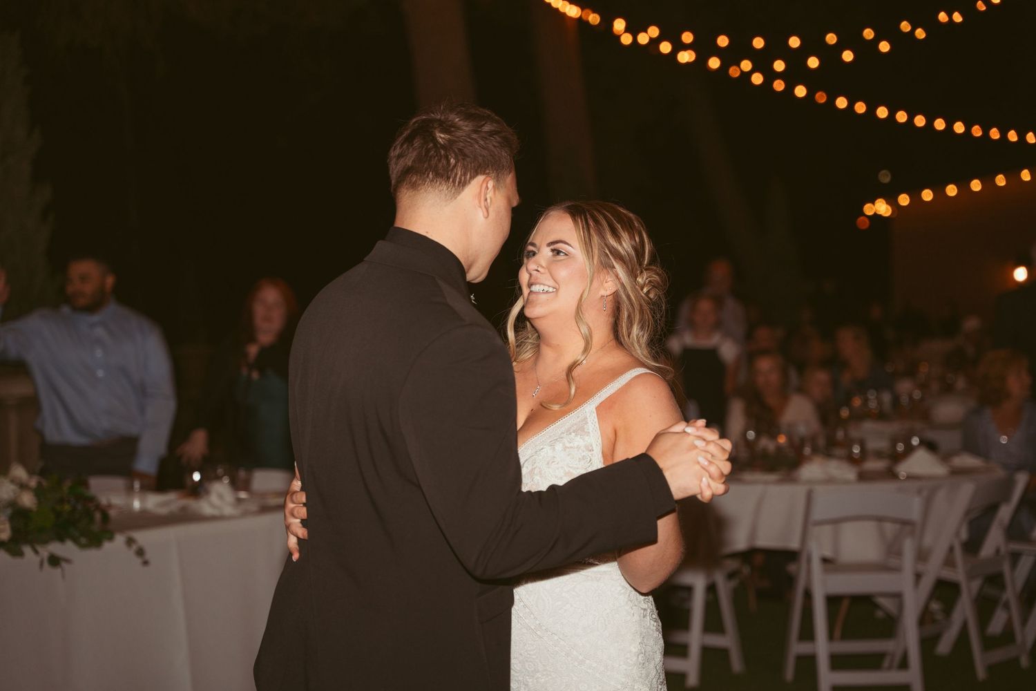 Romantic couple dancing under warm string lights at an outdoor evening wedding reception.