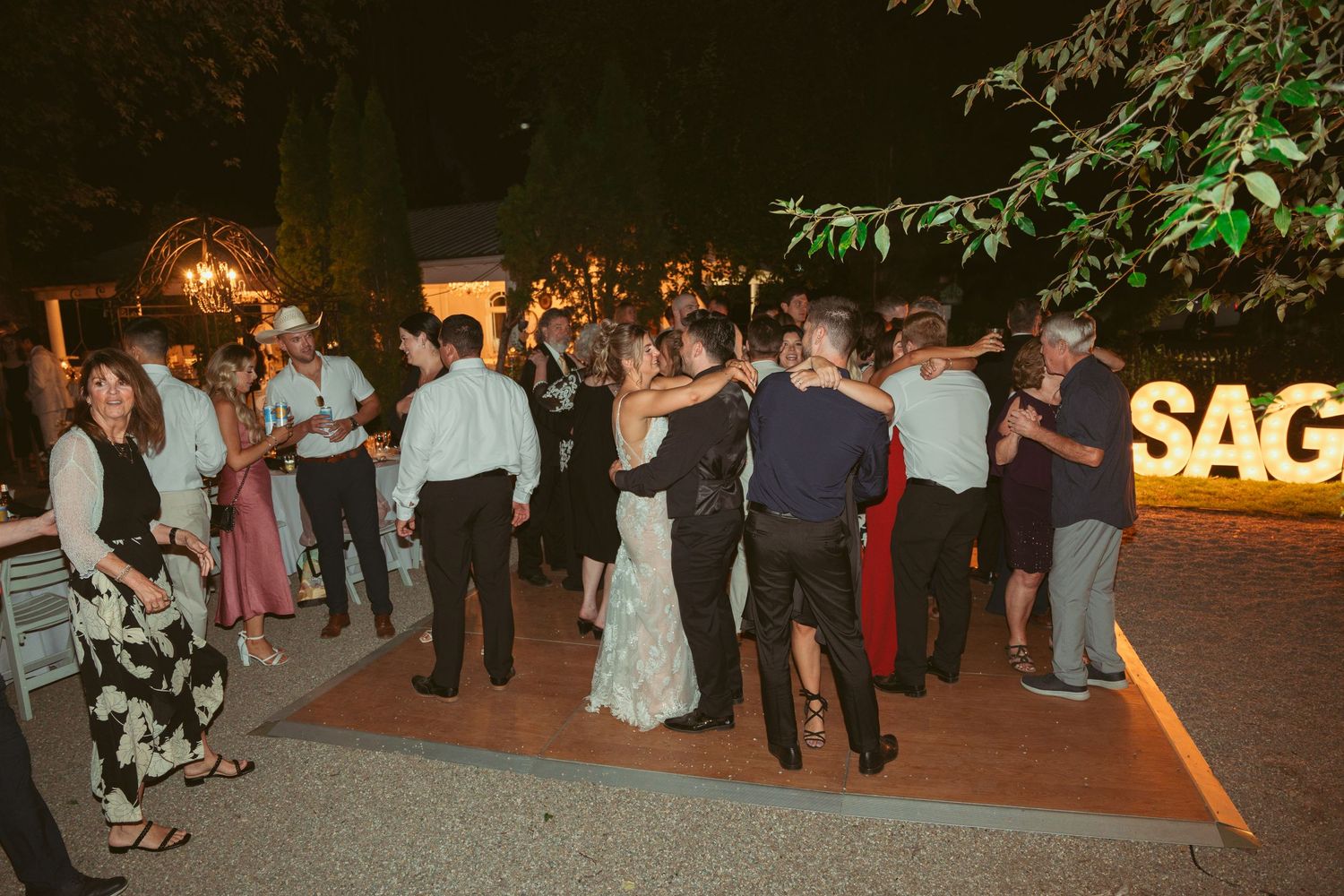 Outdoor wedding celebration at night with illuminated signage and dancing guests.