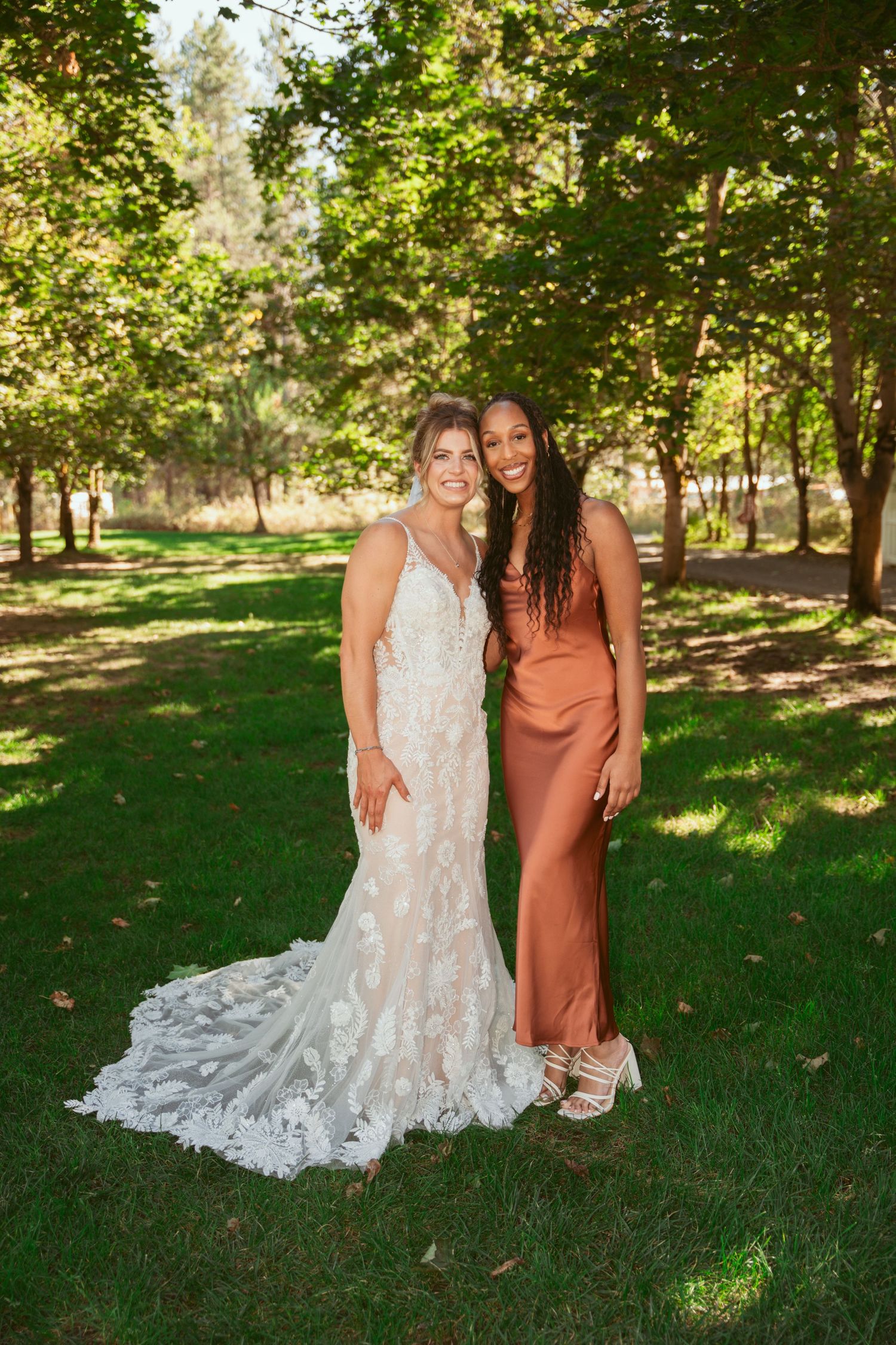 A bride in a lace wedding gown poses with a guest wearing a rust-colored dress on a sunny lawn.