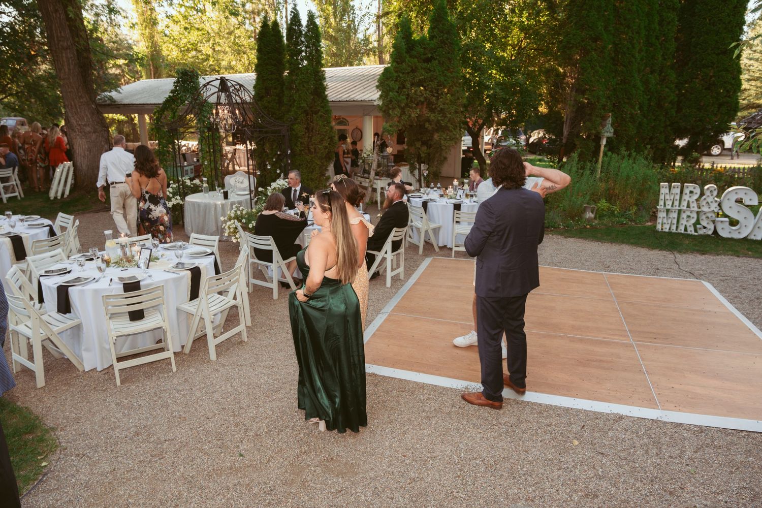 Outdoor wedding reception setup with white tables and dance floor under trees.