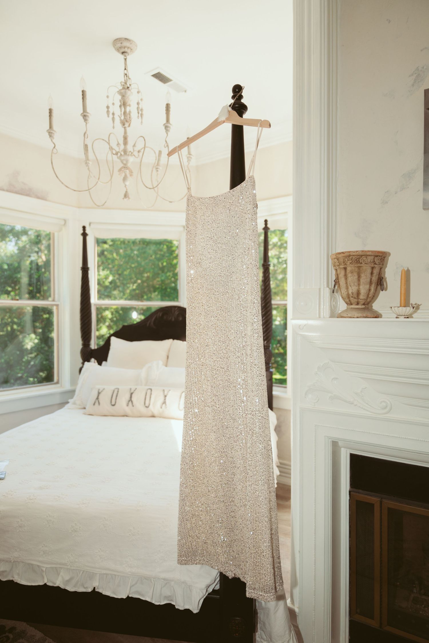 A sparkly white wedding dress hangs elegantly beneath a crystal chandelier in a bright bedroom with a fireplace.