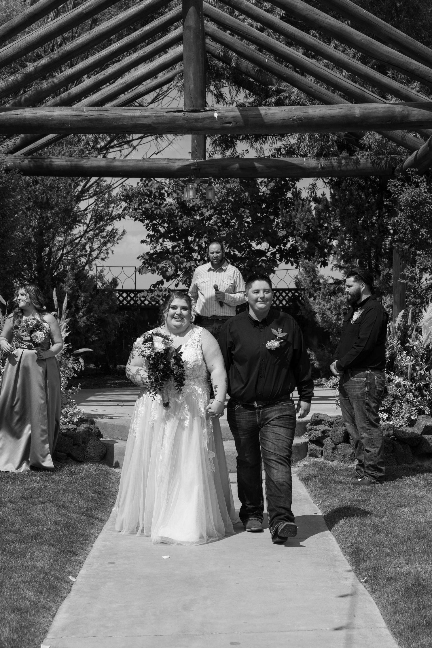 Black and white photo of wedding ceremony under wooden gazebo with guests gathered around.