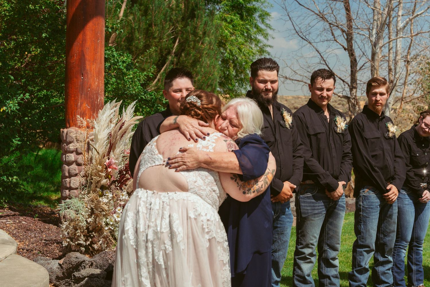Wedding party members pose together outdoors against a natural backdrop with trees and greenery.