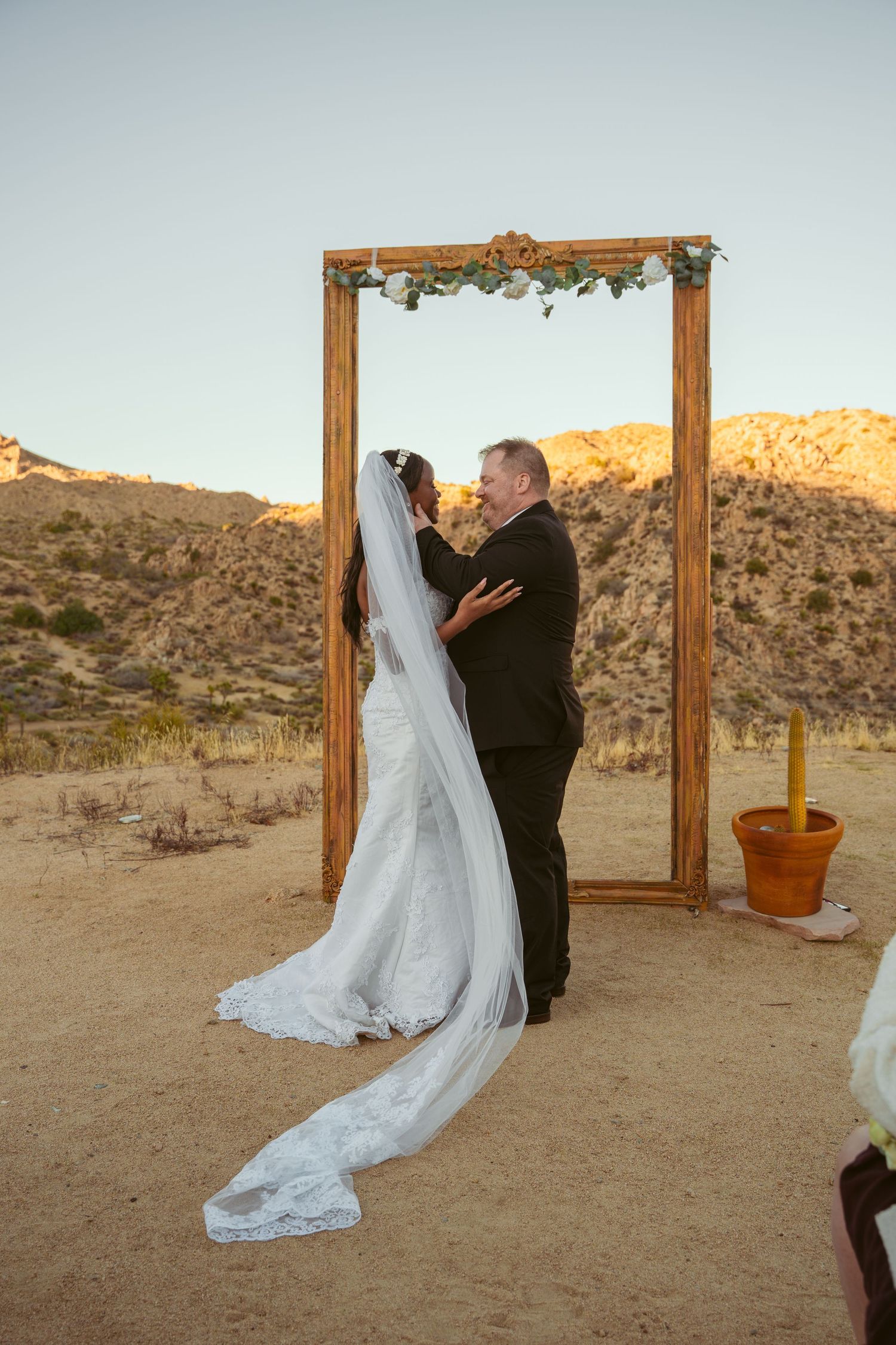 A couple celebrates their wedding moment under a rustic arch with desert mountains glowing at sunset.